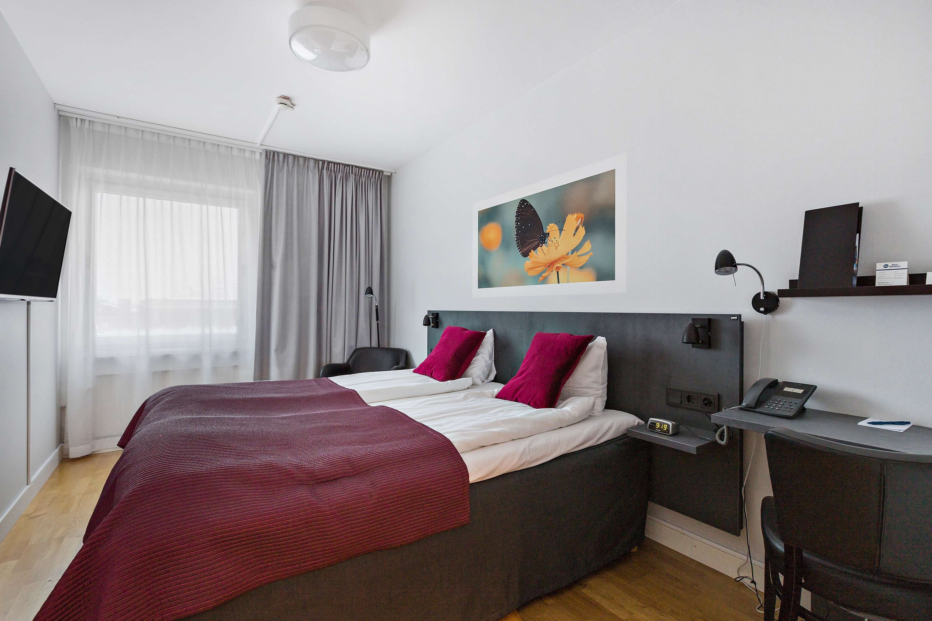 A modern hotel room with a large bed, a desk with a phone, a TV, and a painting on the wall.
