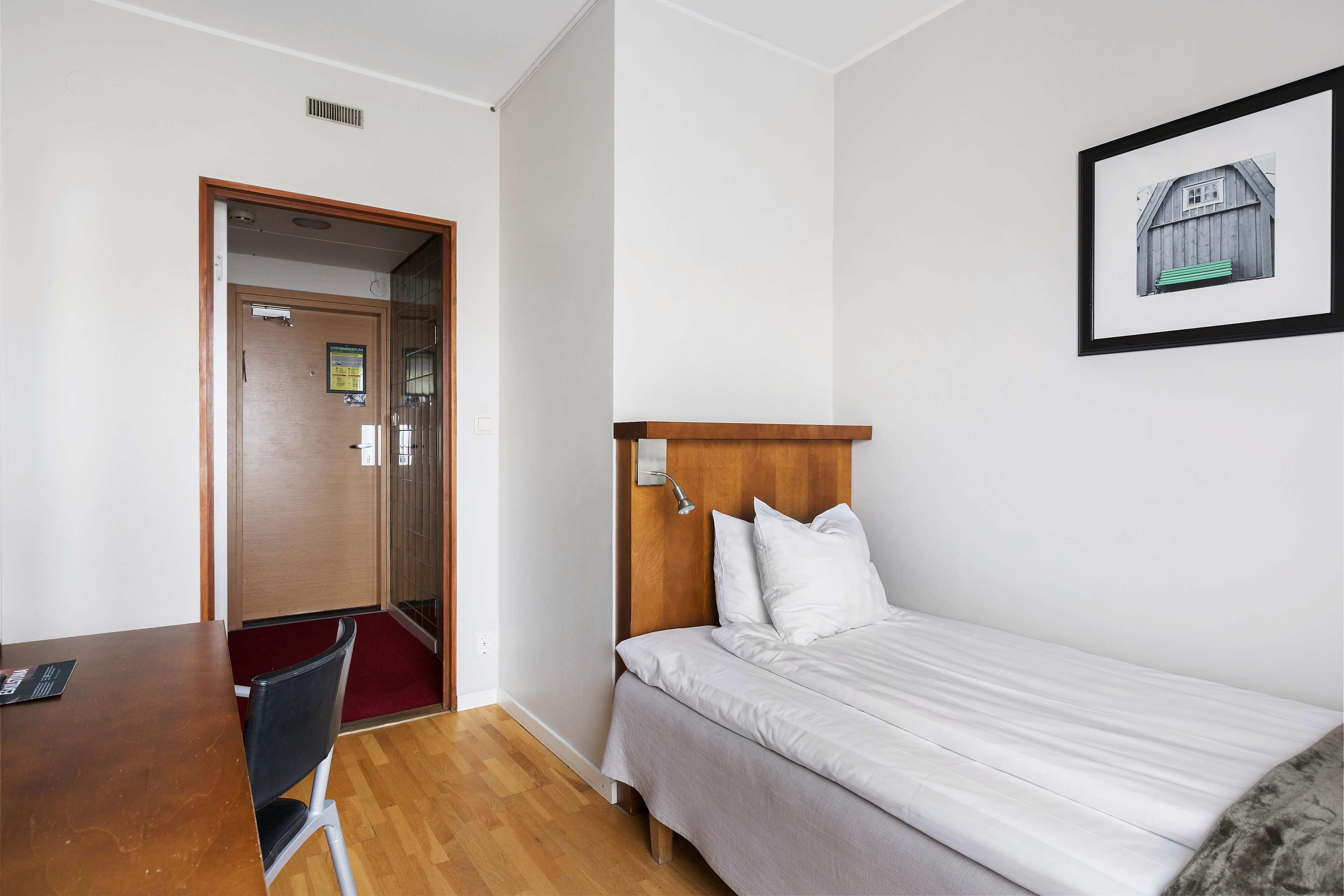 A hotel room with a wooden headboard bed, a desk with a chair, and a door leading to another room.