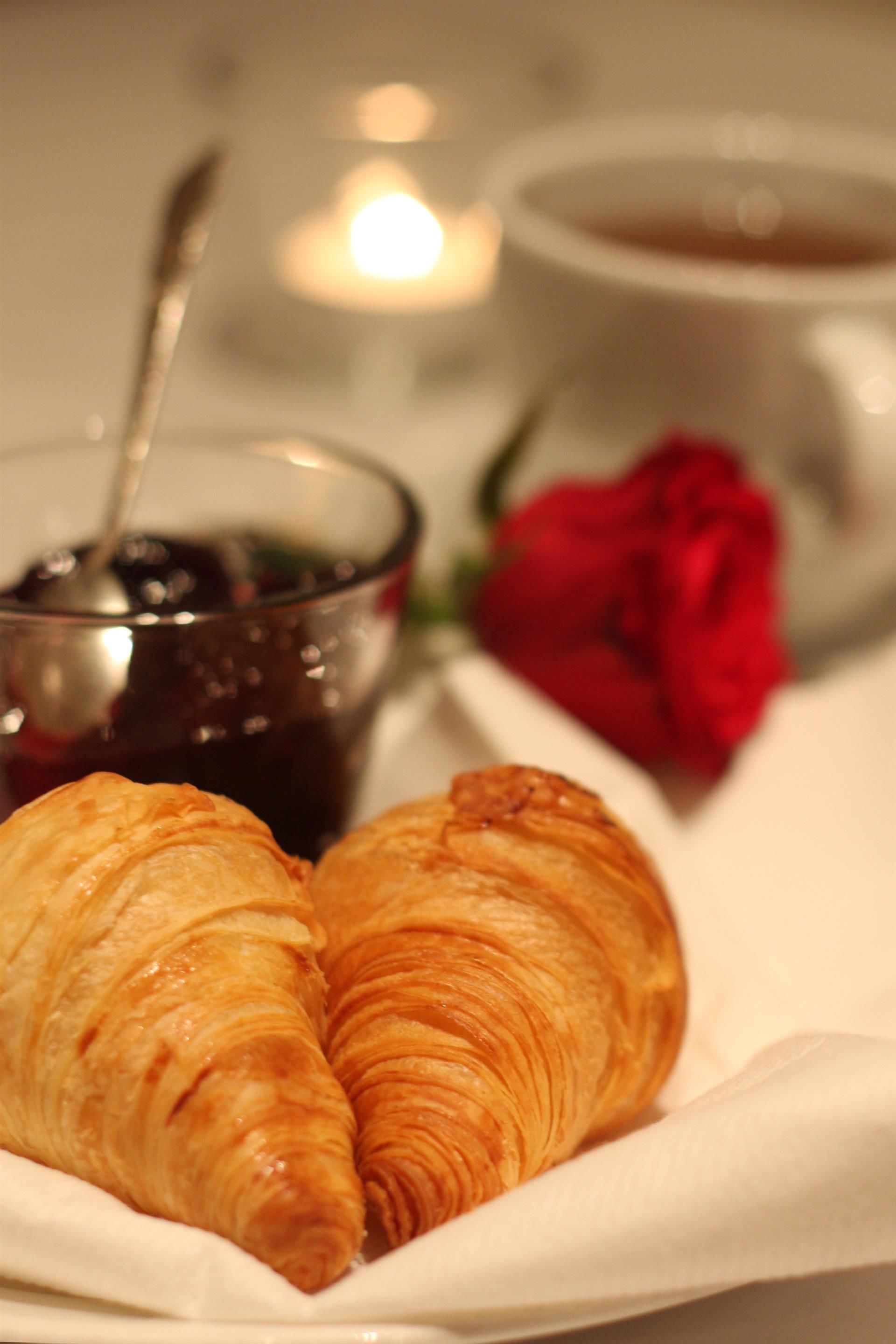 A croissant on a napkin, a cup of coffee, a glass of jam, and a red rose.