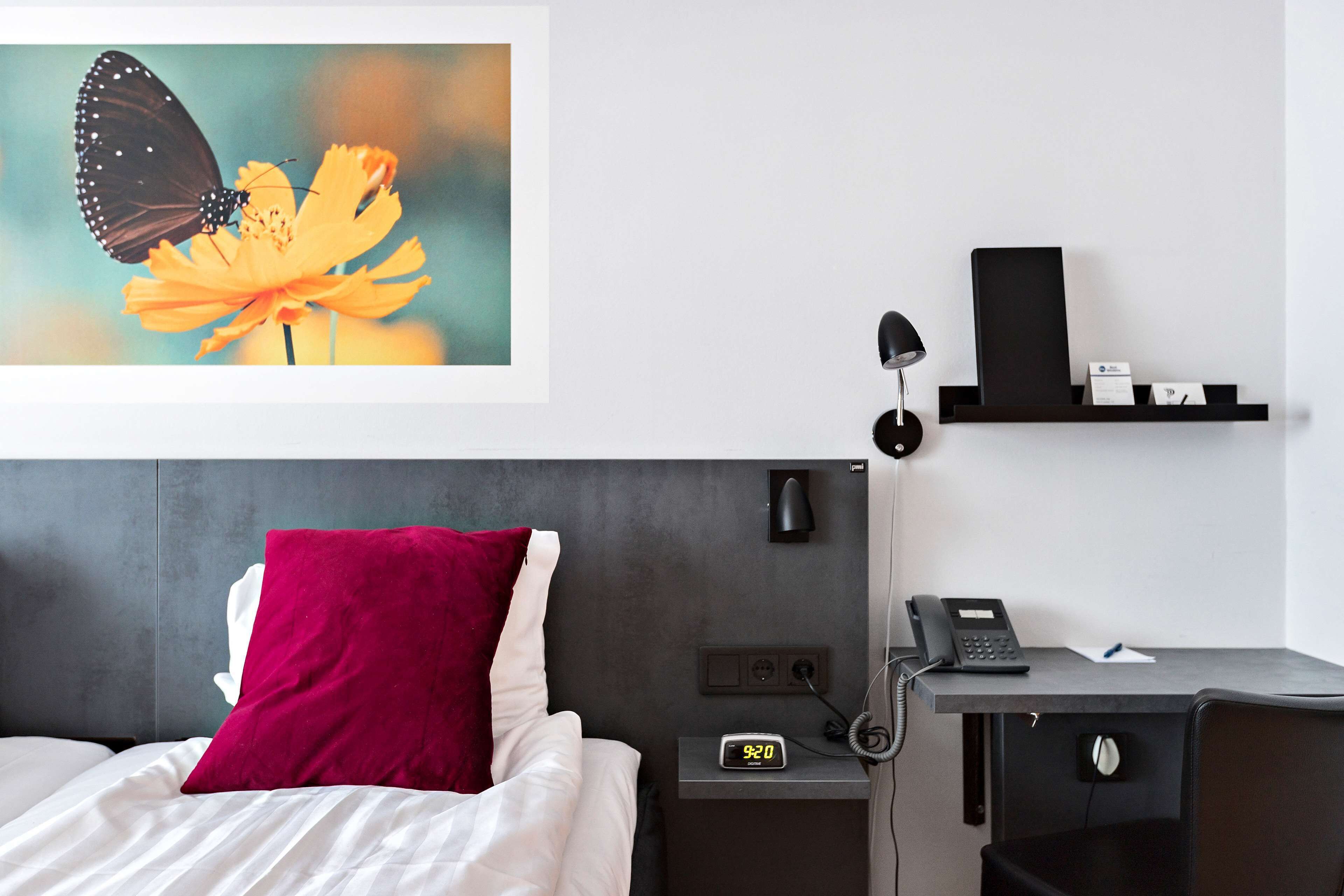 A hotel room with a bed, a nightstand, a desk, a phone, a clock, and a picture of a butterfly on a flower.