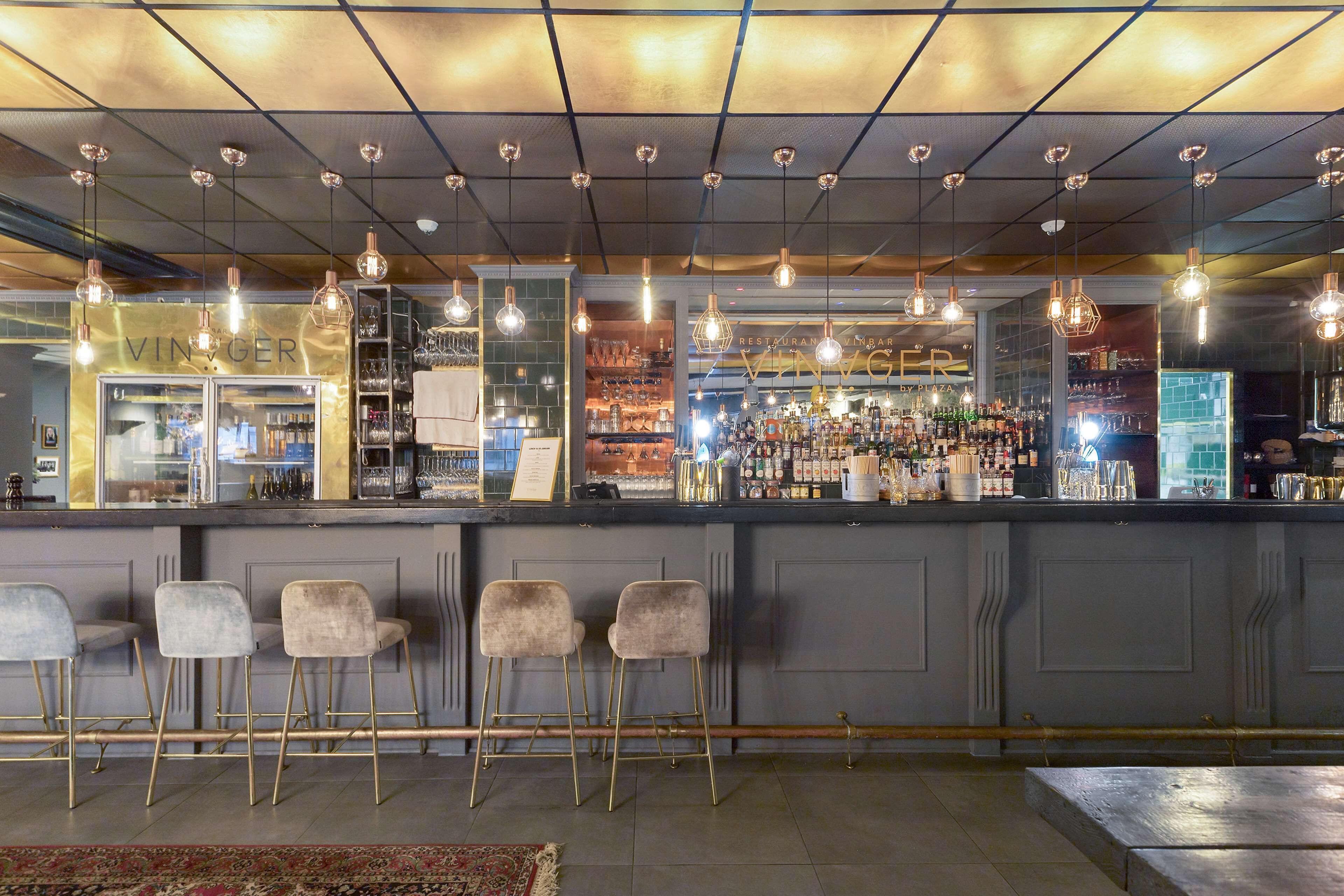 Una zona de bar con mostrador, taburetes, lámparas colgantes y estantes llenos de botellas y vasos.