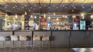 Una zona de bar con mostrador, taburetes, lámparas colgantes y estantes llenos de botellas y vasos.