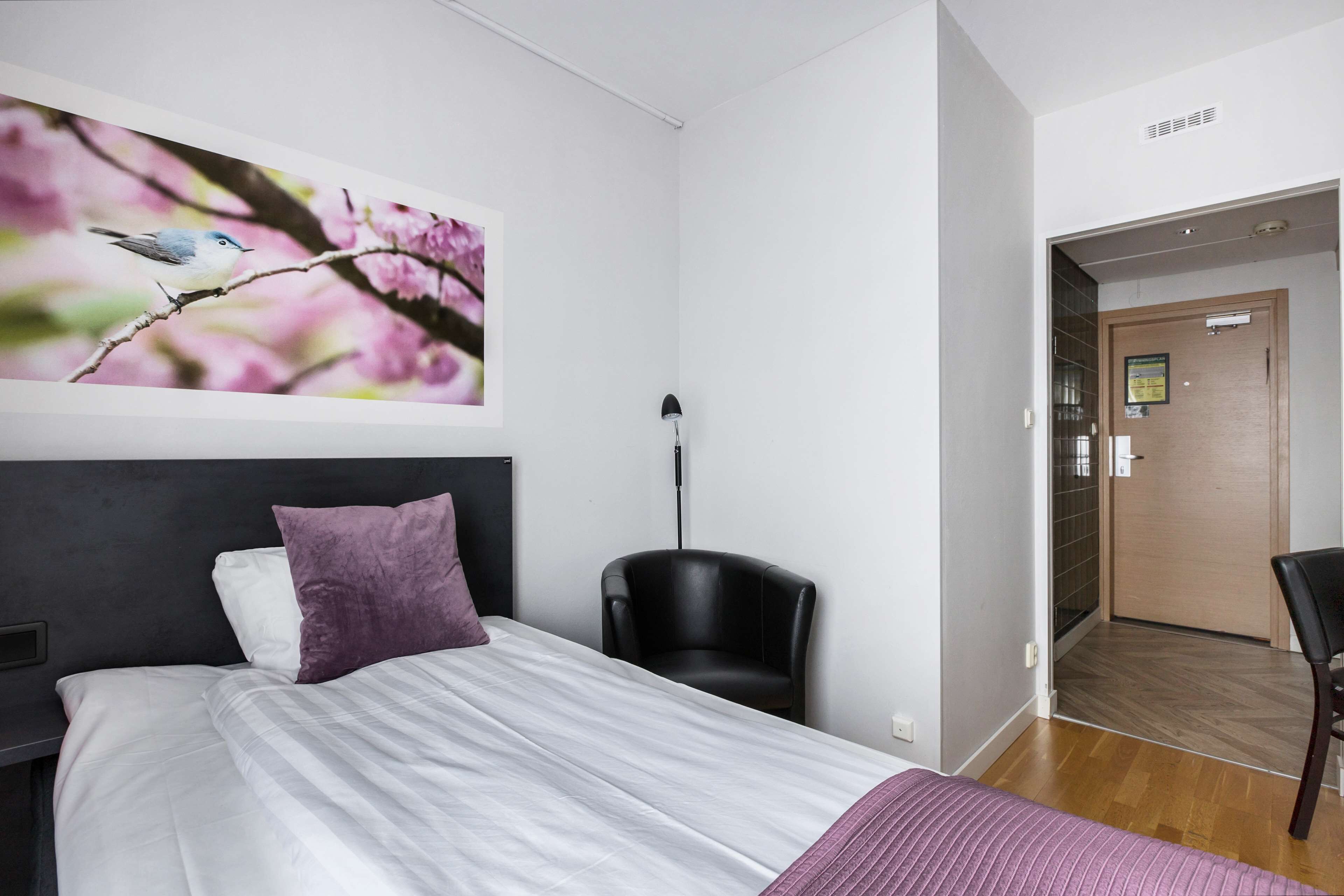 Ett modernt hotellrum med en stor säng, en stol och en dörr som leder ut till en korridor.