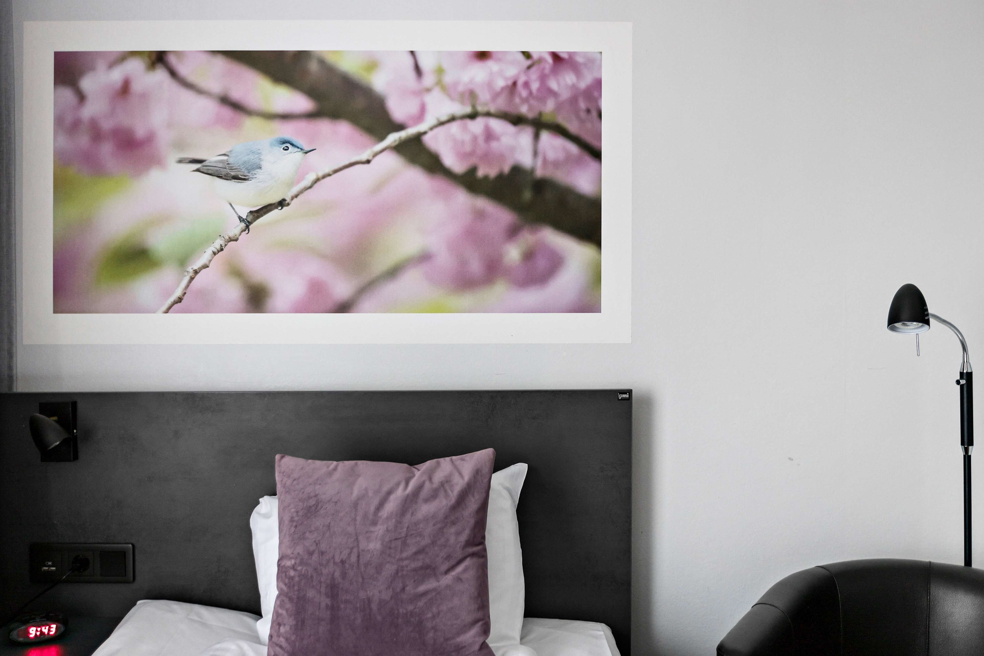 A bedroom with a bed, a nightstand, a lamp, and a wall-mounted picture of a bird on a branch.