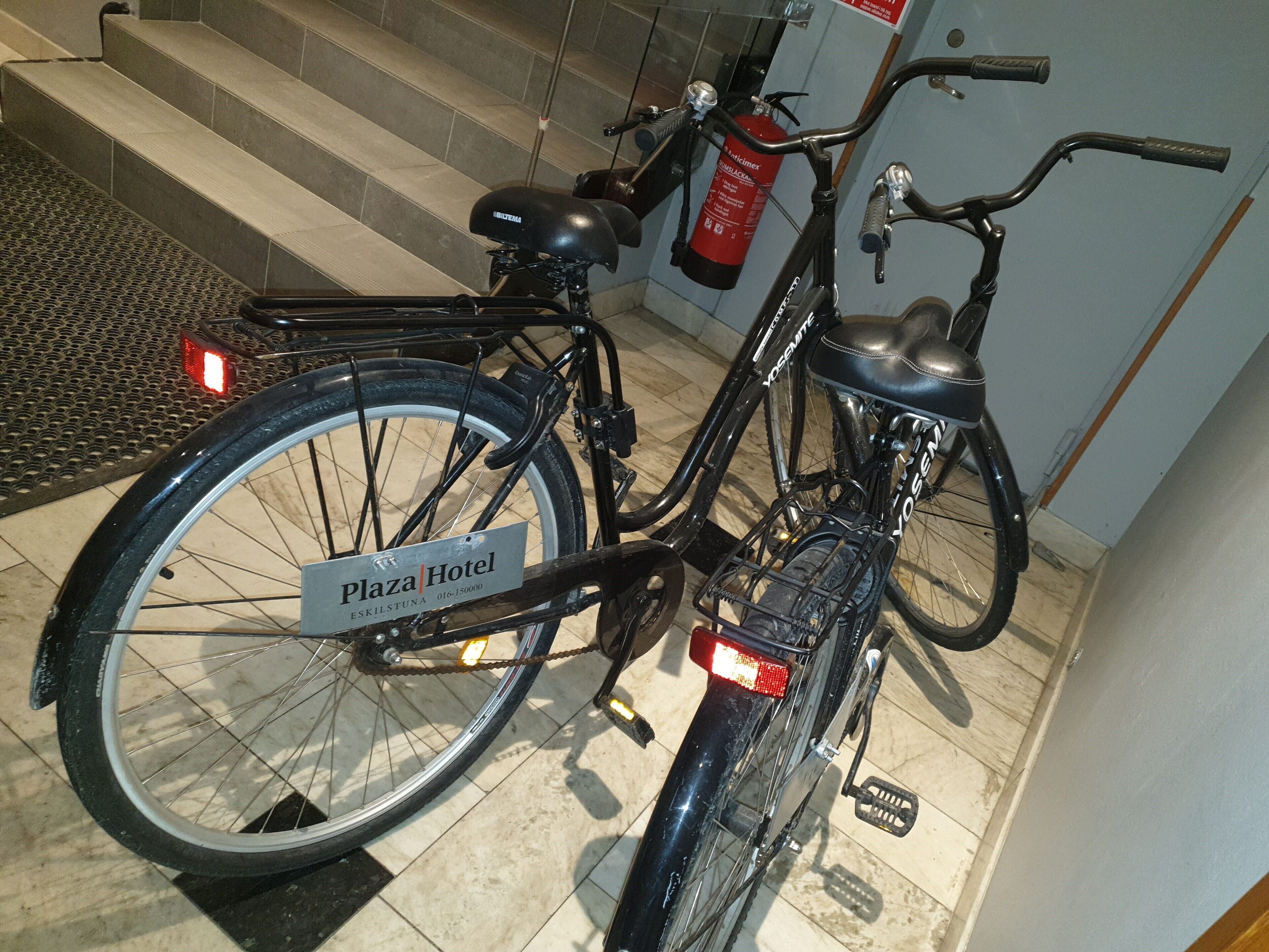 A bicycle with a 'Plaza Hotel' sign attached to the frame, parked indoors near a staircase.