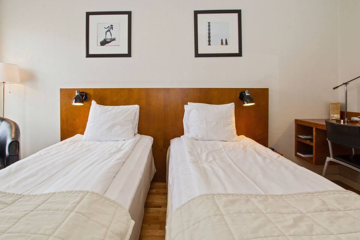 Een hotelkamer met twee bedden, een bureau, een stoel en twee ingelijste foto’s aan de muur.