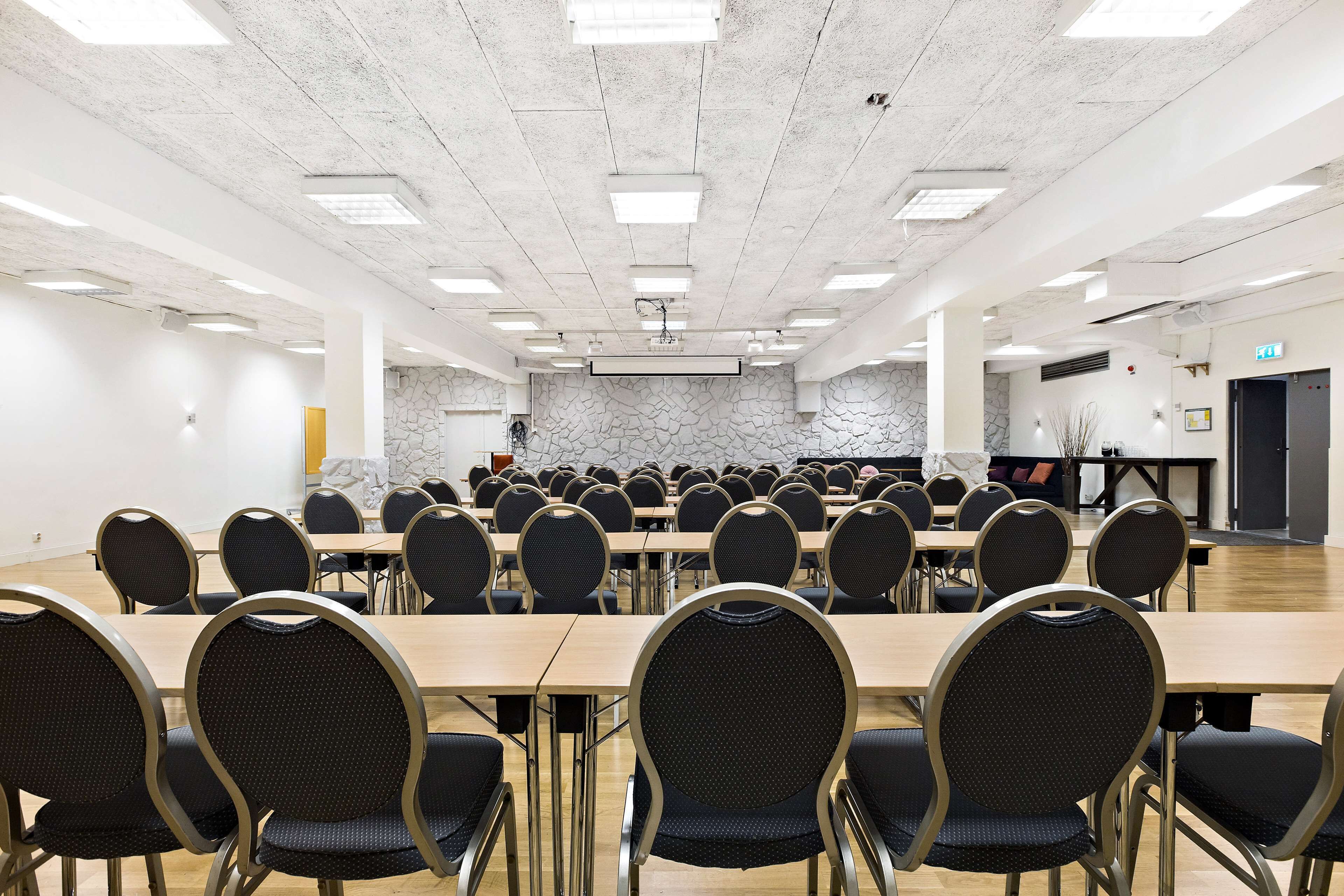 A conference room with multiple tables and chairs, a stone wall, and a projector screen.