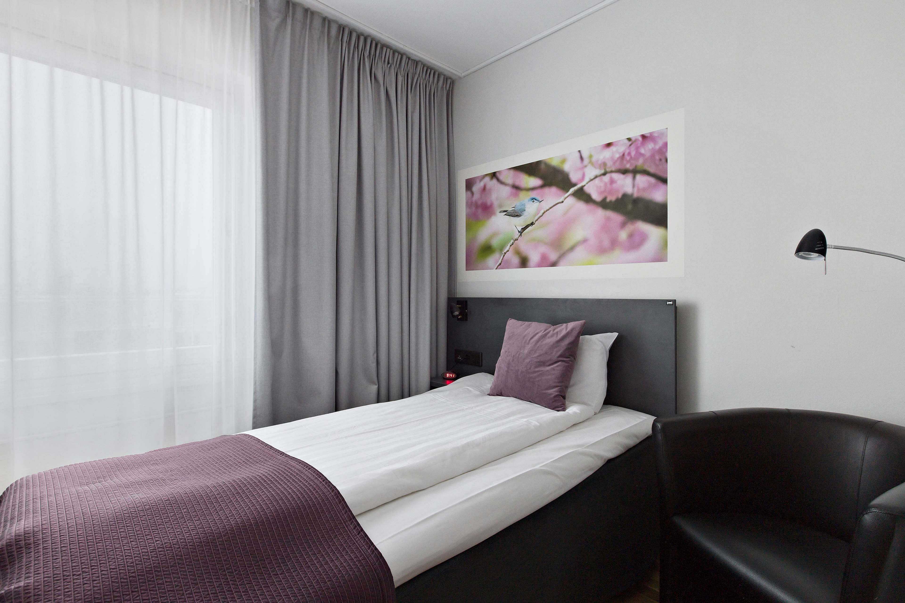 A modern hotel room with a bed, a chair, a bedside table, a lamp, and a framed picture on the wall.