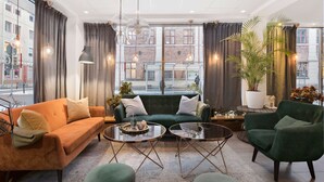 A modern living room with a green sofa, orange armchair, and a green armchair, featuring a glass coffee table, pendant lights, and large windows with curtains. - Best Western Plaza Hotel (Eskilstuna)