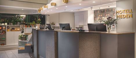 A hotel reception desk with a modern design, a visible name sign, and a variety of amenities including a plant display and a small seating area.