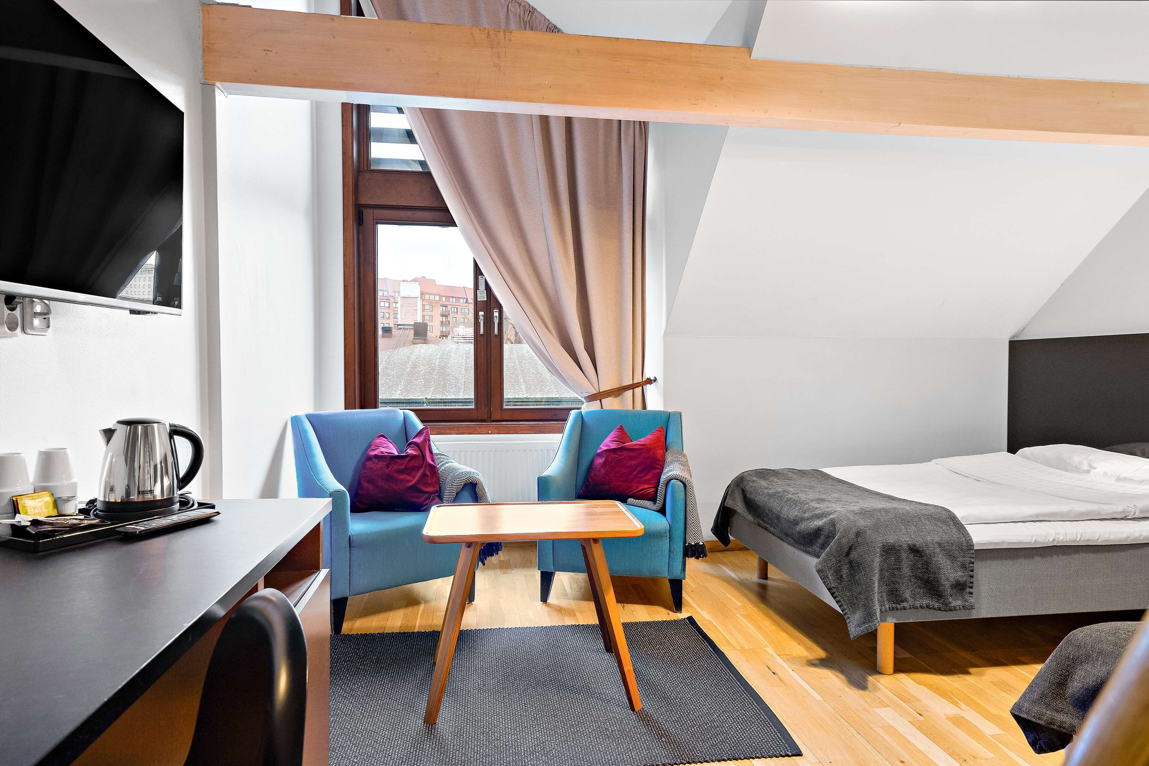 A modern room with a bed, two blue armchairs, a small wooden table, a desk with a kettle, and a window with curtains.