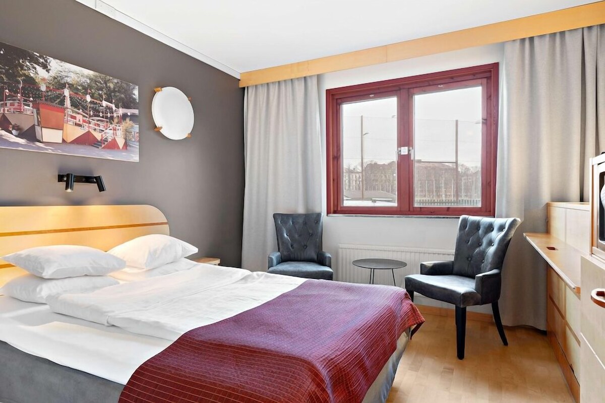 A hotel room with a bed, two armchairs, a small table, a window with curtains, a wall-mounted round mirror, and a picture on the wall.