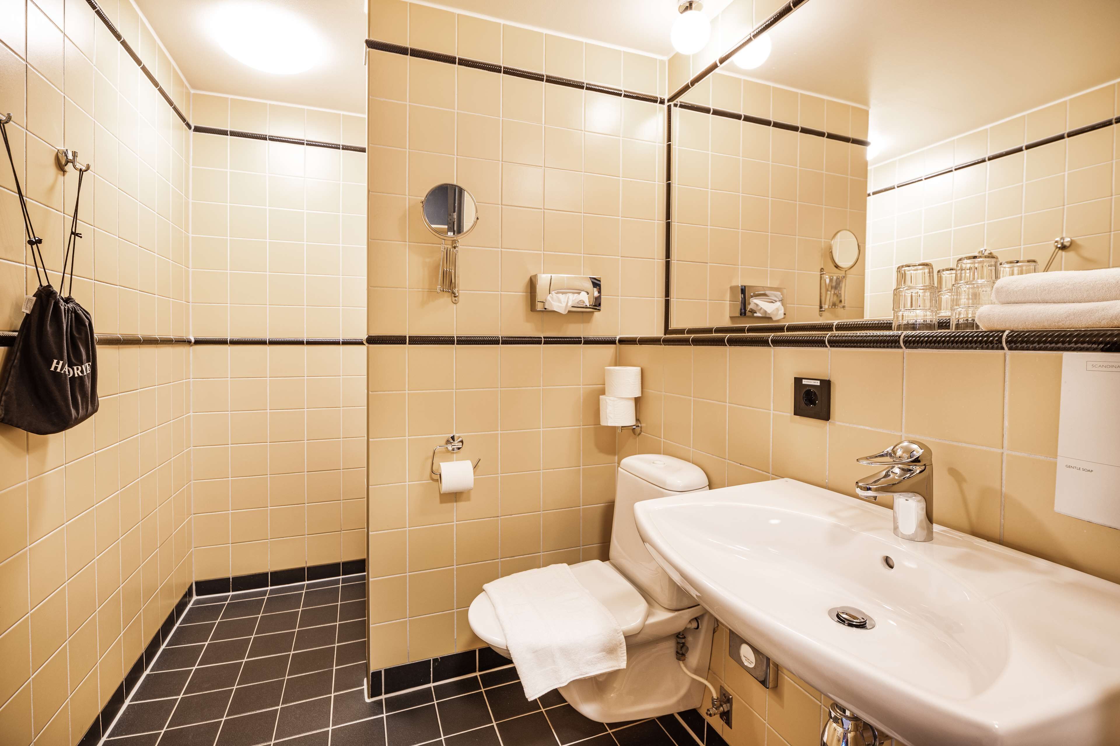 A bathroom with a white sink, mirror, and toilet. There is a towel hanging on the wall and a bag hanging on a hook.