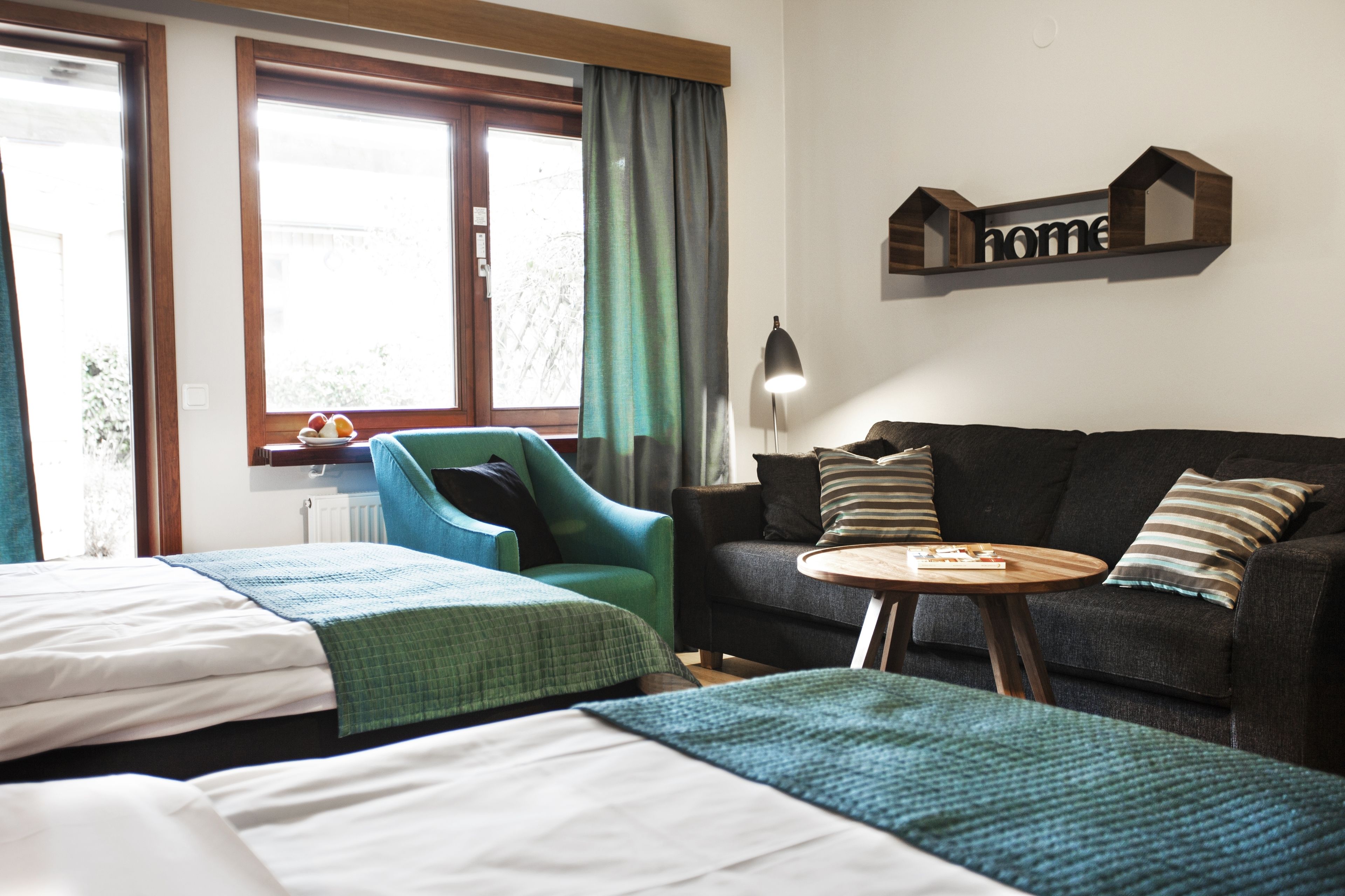 Habitación de hotel con dos camas, un sillón verde, un sofá oscuro, una mesa redonda de madera y una lámpara.