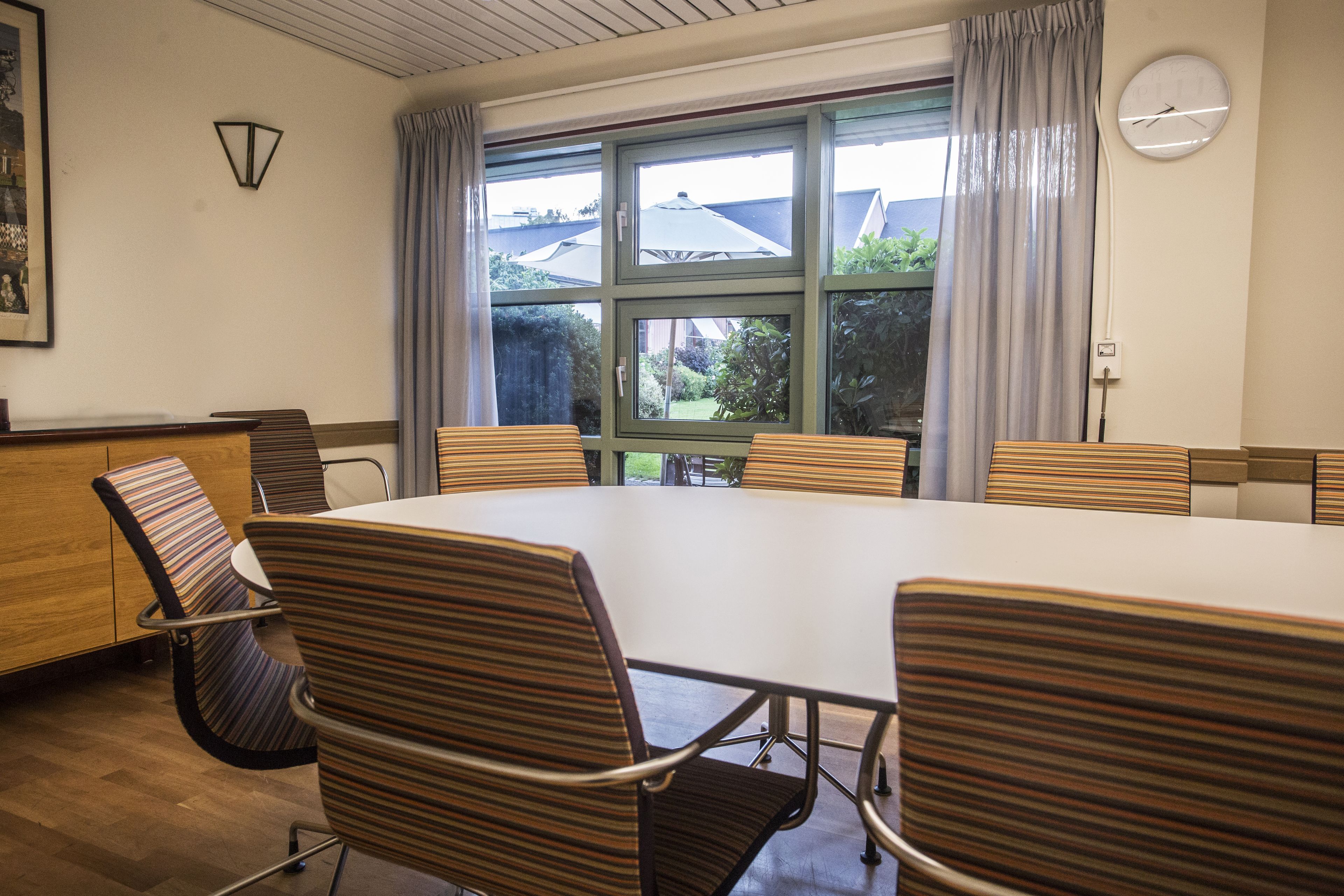 Un salón de conferencias con una gran mesa y seis sillas a rayas, un reloj en la pared y una ventana con cortinas.