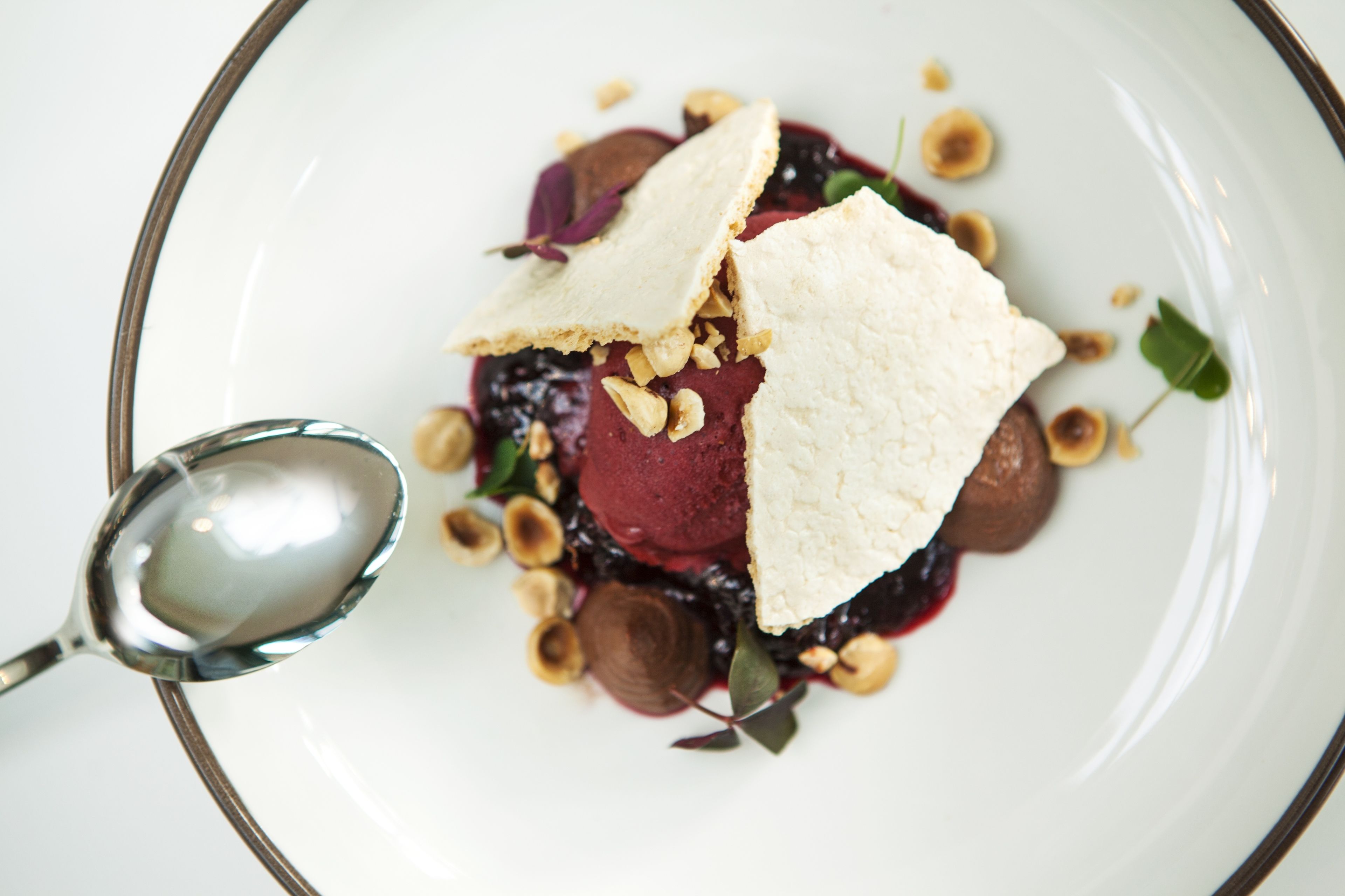 A white plate with a dessert consisting of a scoop of ice cream, a piece of cake, and nuts, accompanied by a spoon.