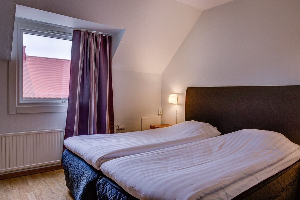 A bedroom with two beds, a window with curtains, a bedside lamp, and a radiator.
