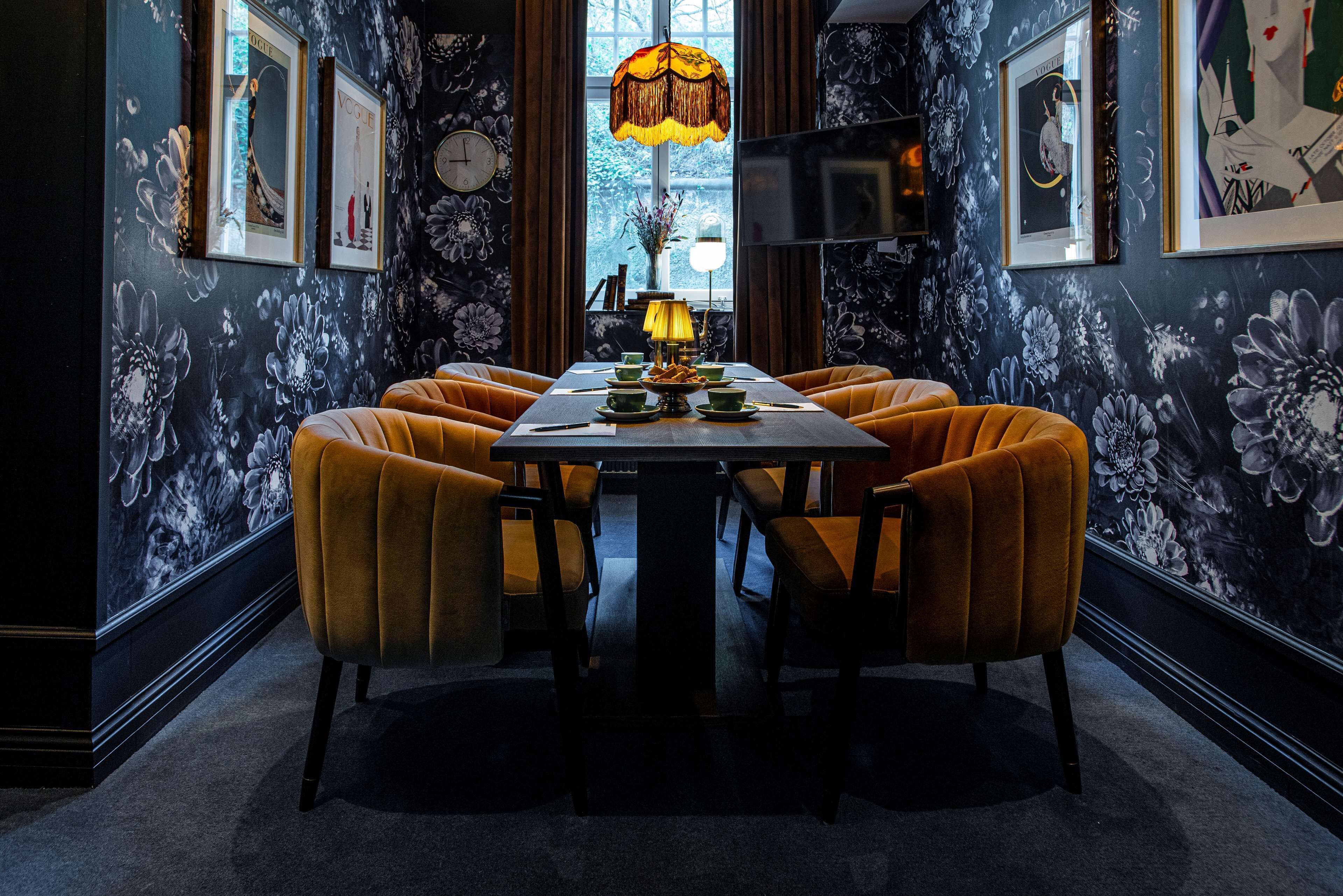 A dining area with a long table, six chairs, a chandelier, and floral wallpaper.