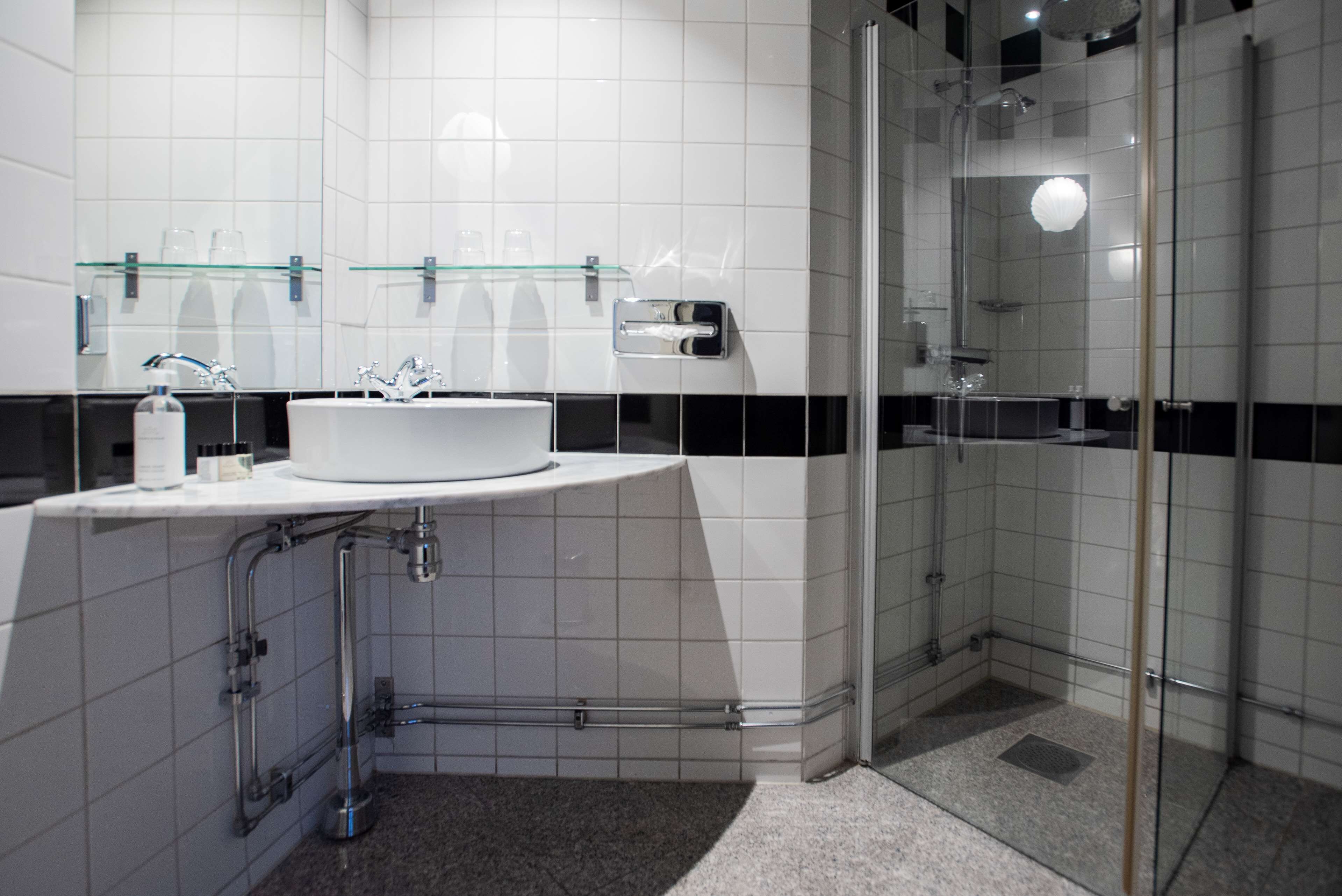 A modern bathroom with a glass shower enclosure, a white sink with a glass shelf above, and a hand dryer.