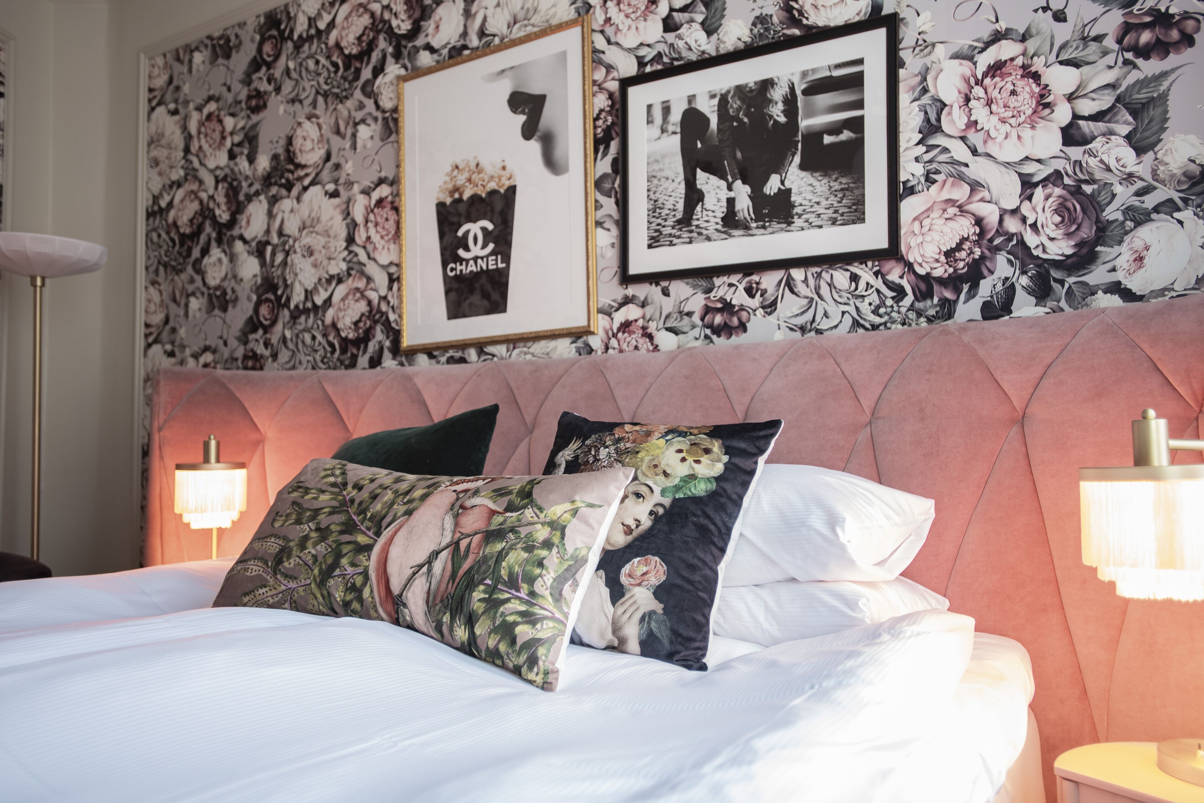 A neatly made bed with floral and Chanel-themed pillows, a tufted headboard, and framed pictures on the wall.