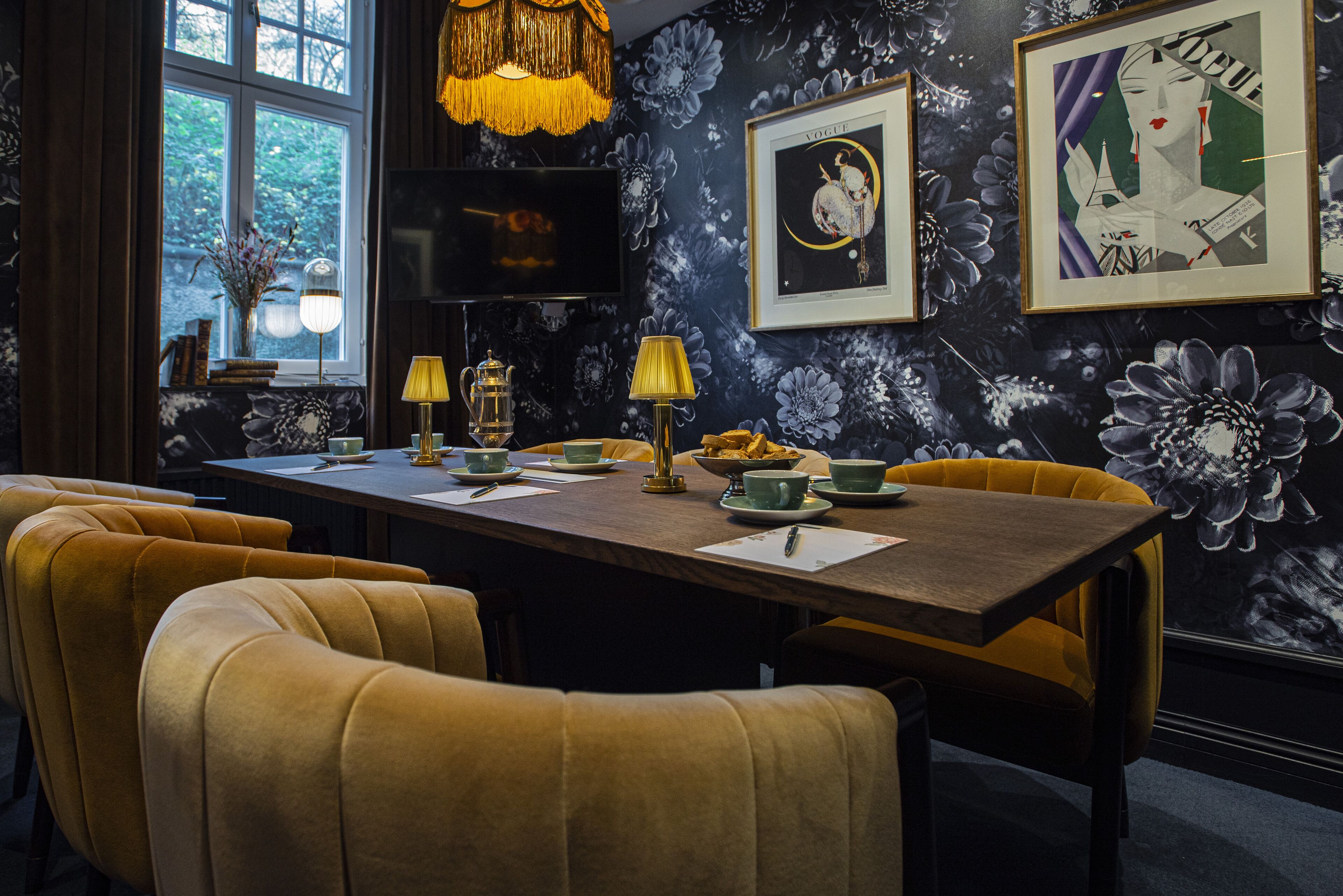 Un comedor con papel tapiz floral oscuro, una mesa de madera, sillas amarillas y una araña dorada.