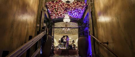 Una gran escalera con techo floral y candelabro, que conduce a un comedor con mesas y sillas.