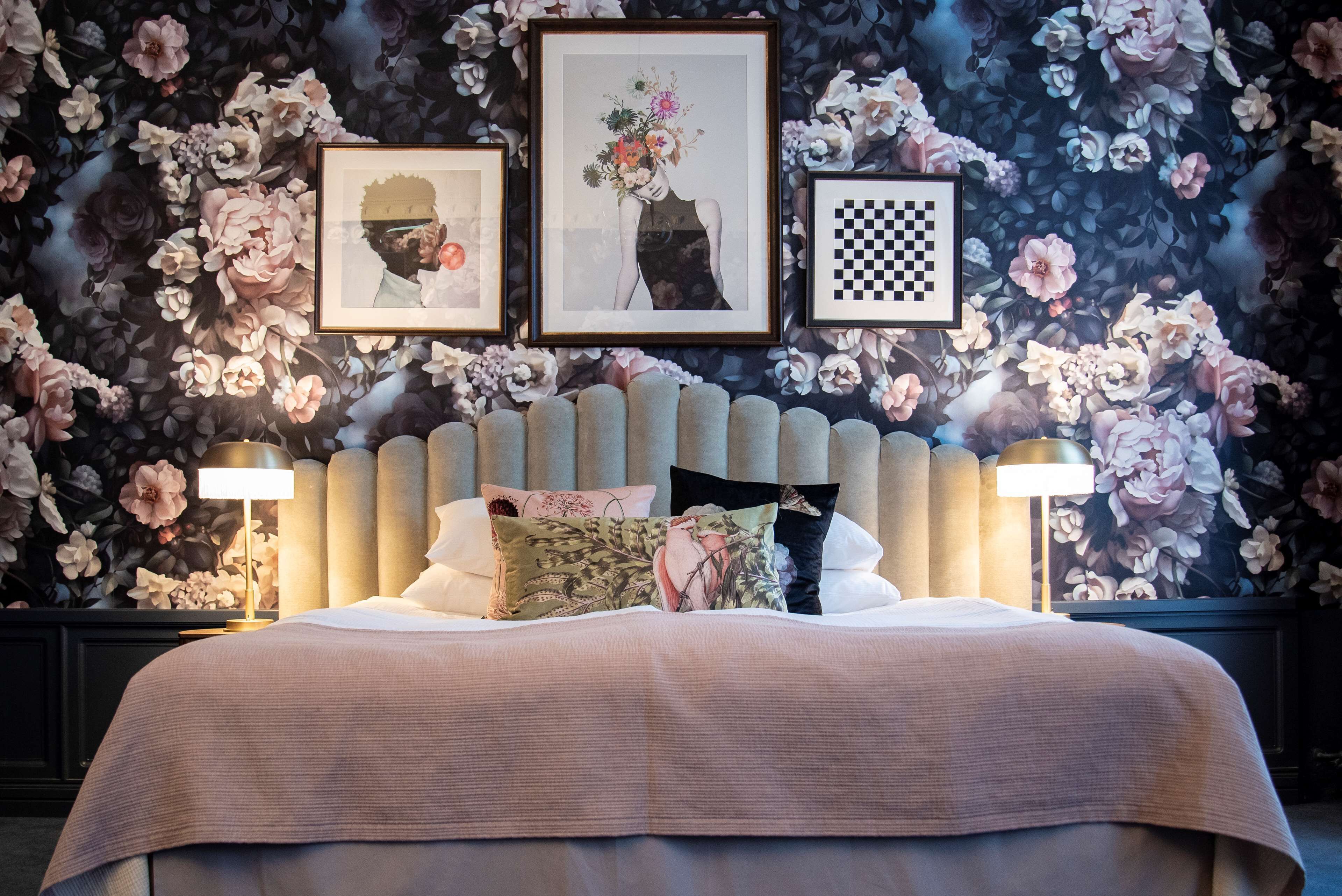 A bedroom with a large bed, two bedside lamps, a floral wallpaper, and three framed pictures on the wall.