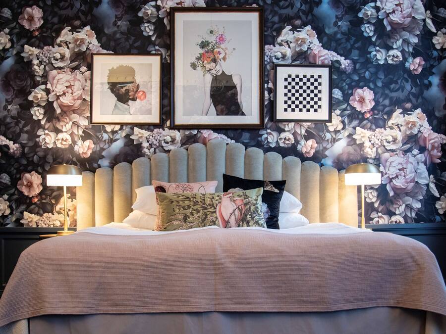 A bedroom with a large bed, two bedside lamps, a floral wallpaper, and three framed pictures on the wall.
