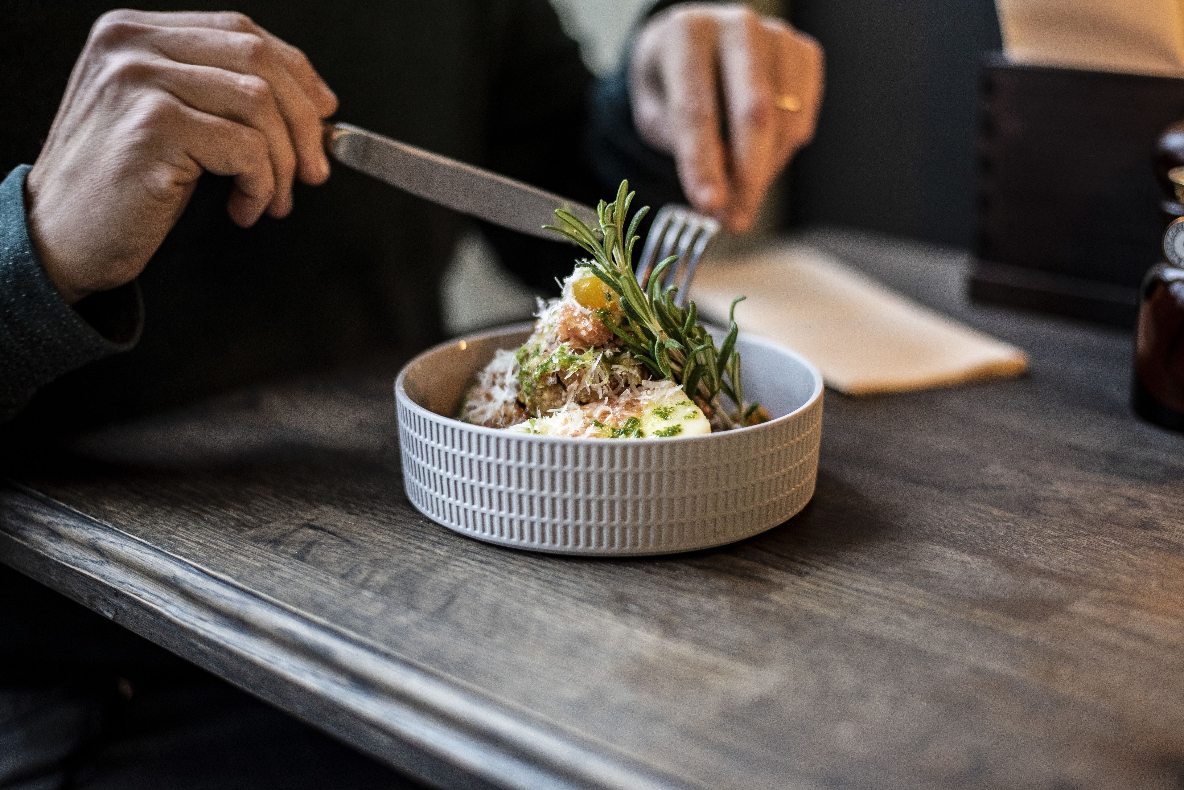 A person holding a knife and fork, preparing to eat from a bowl on a wooden table.