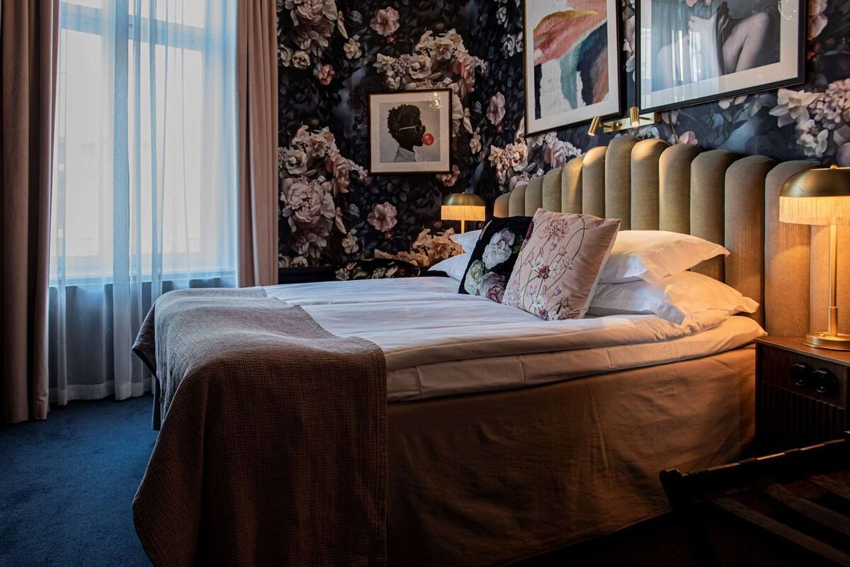 Hotellihuone, jossa on suuri sänky, kukallinen tapetti, sängynpääty, yöpöytä lampulla ja ikkuna, jossa on verhot.