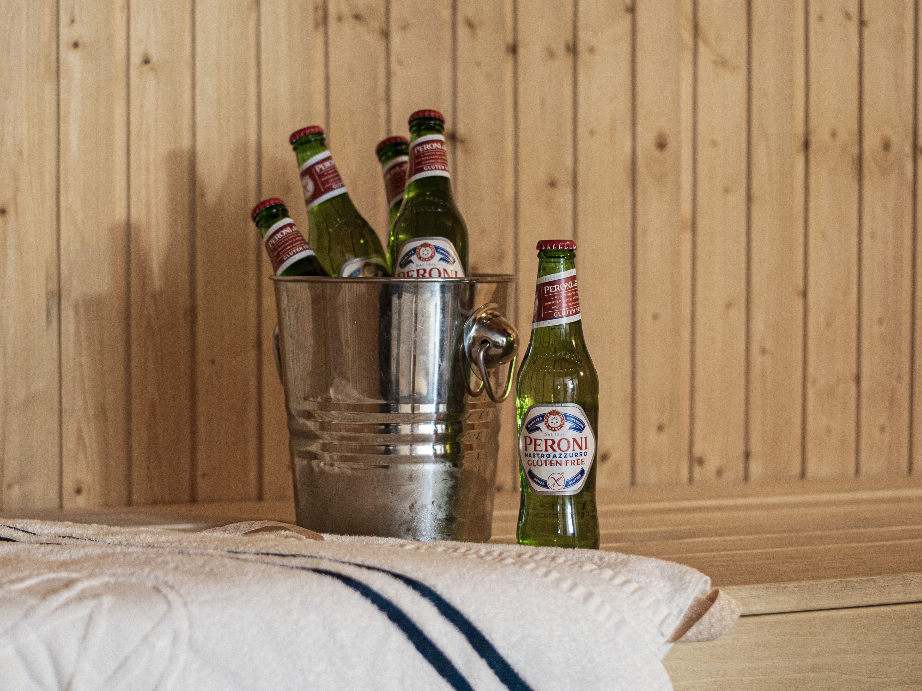 A metal bucket filled with bottles of Peroni beer, placed on a wooden surface with a towel in front.