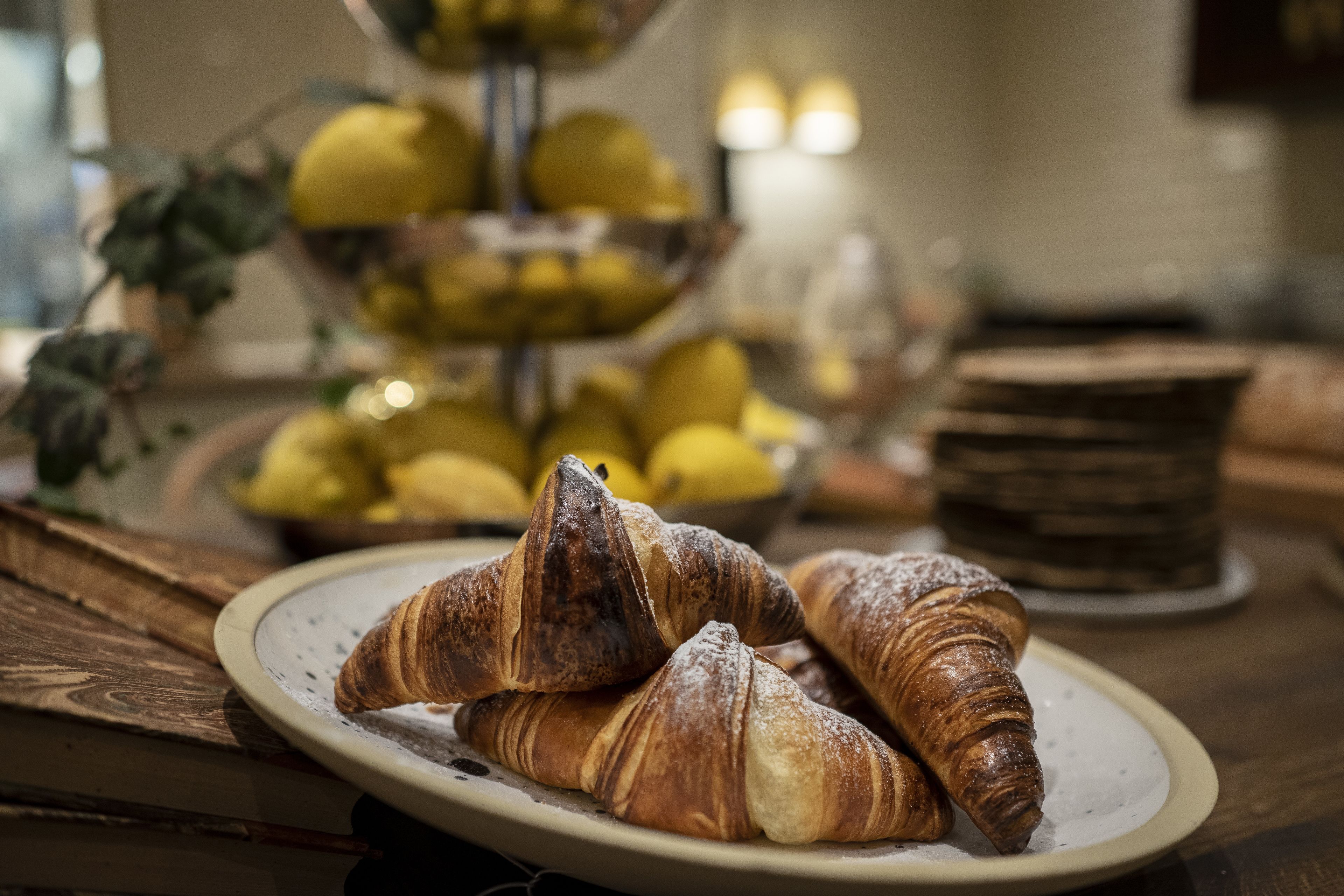 A plate with croissants on a wooden table, a bowl of lemons, and a stack of bread rolls in the background.
