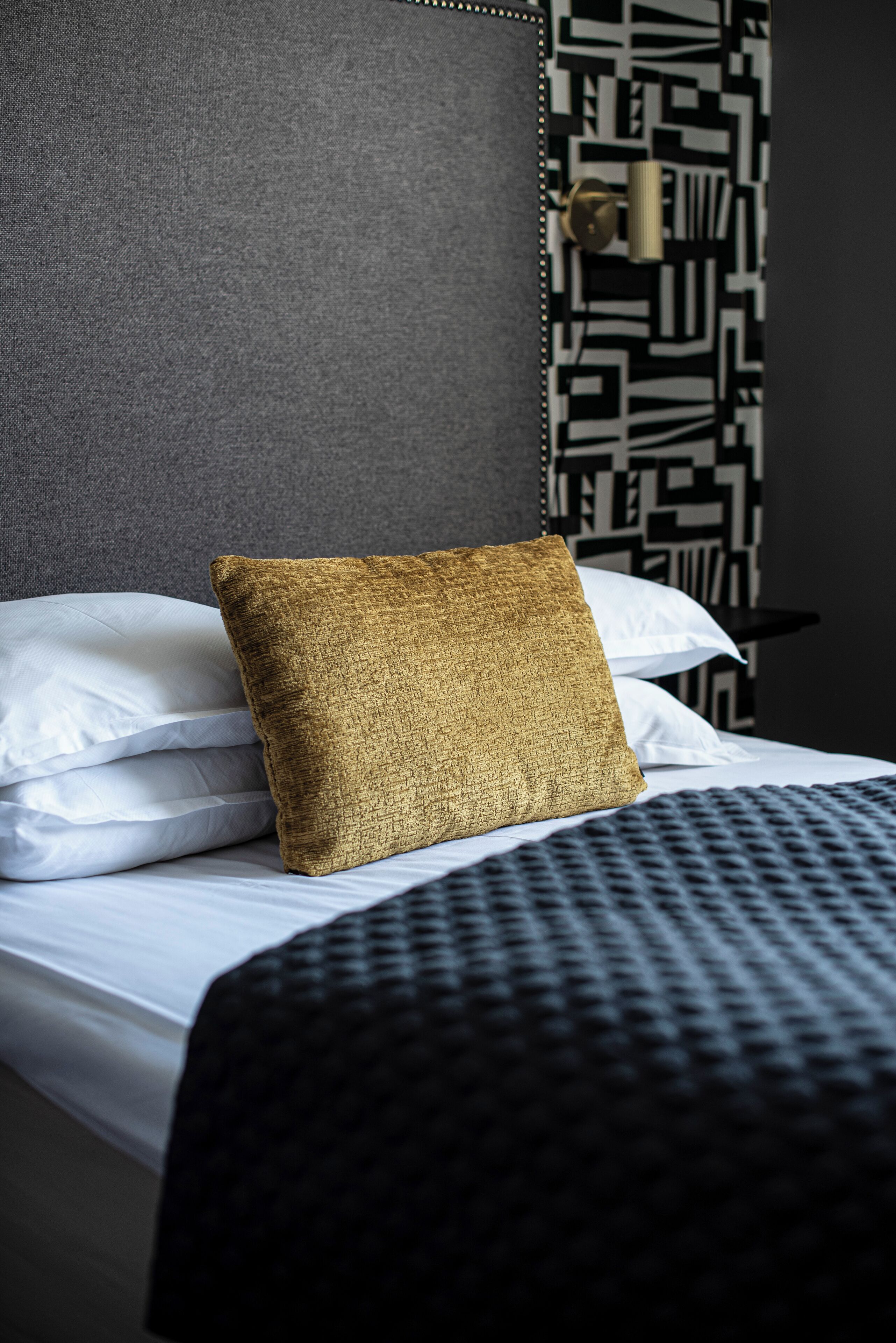A bed with white pillows and a textured gold pillow, a dark headboard, and a patterned wall.