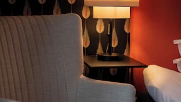 Ein Zimmer mit einem gepolsterten Sessel, einem Nachttisch mit Lampe und einem Bett an einer roten Wand mit einem Wandpapier mit Blattmuster.