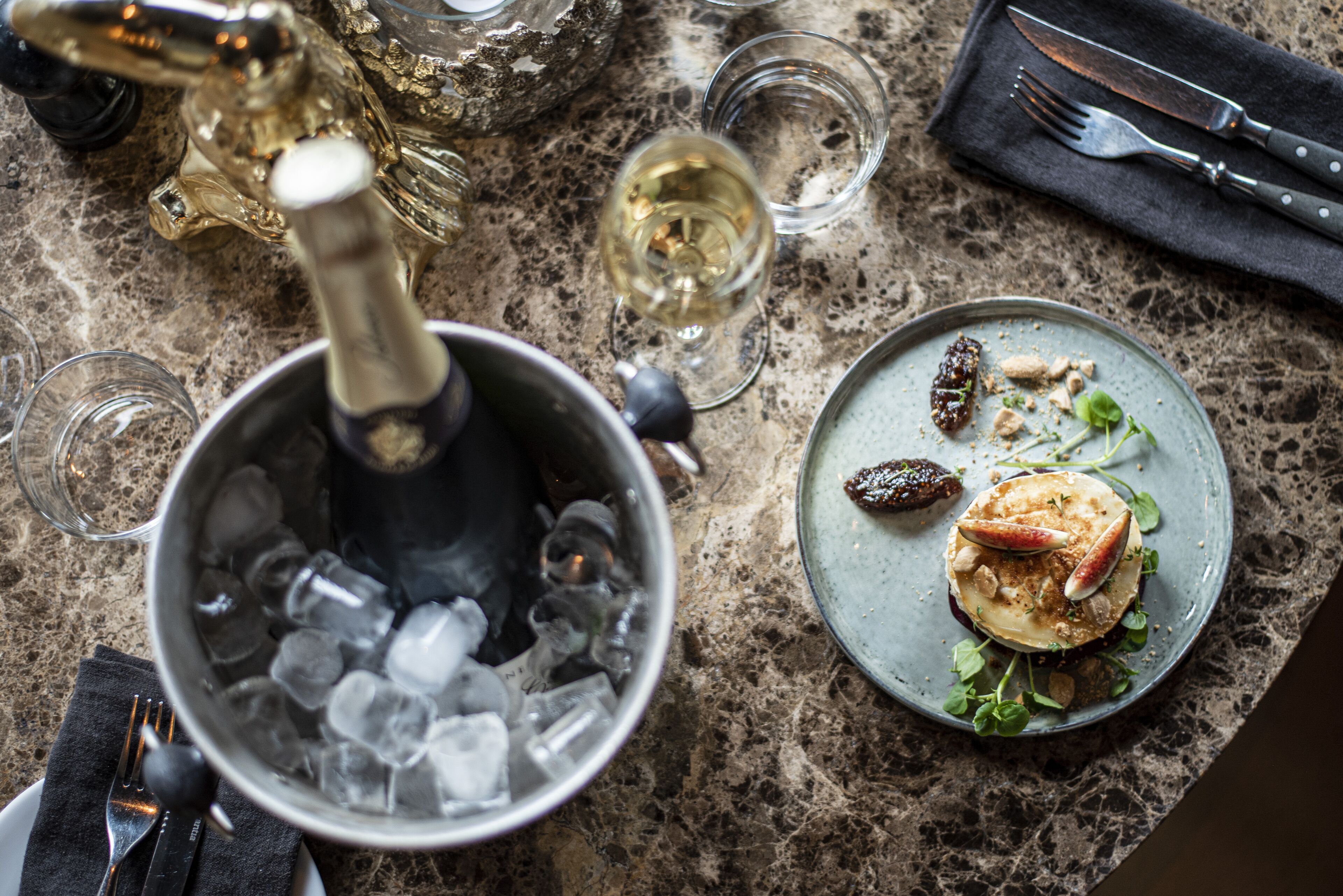 A dining table with a bottle of champagne, a glass of wine, and a plate of food.