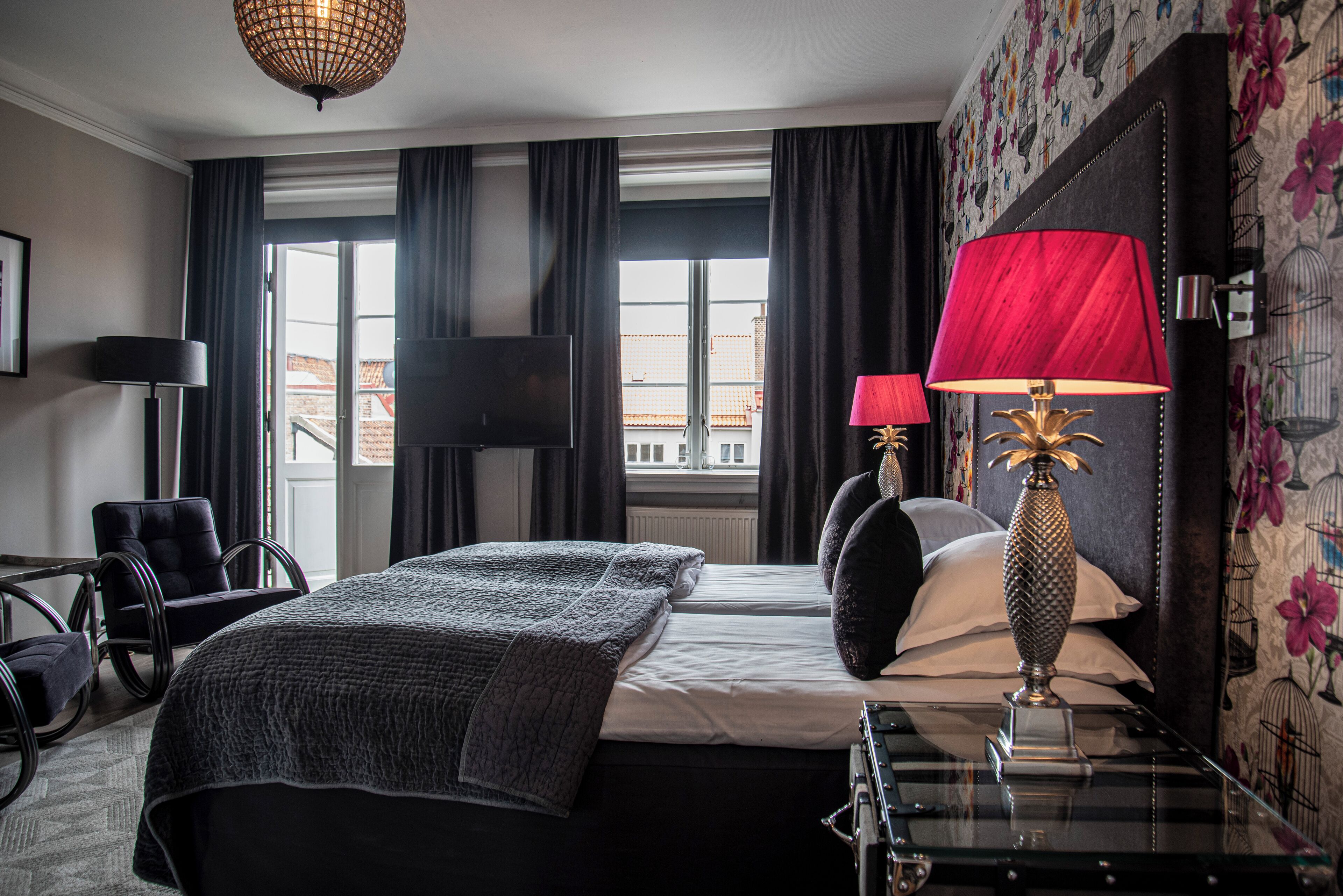 A hotel room with a bed, two bedside lamps, a chair, a TV, and a view of buildings through the window.