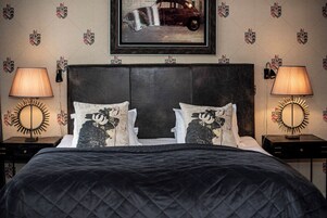 Una cama bien hecha con un edredón oscuro, dos almohadas con diseño de vaca y un cuadro de un coche en la pared.