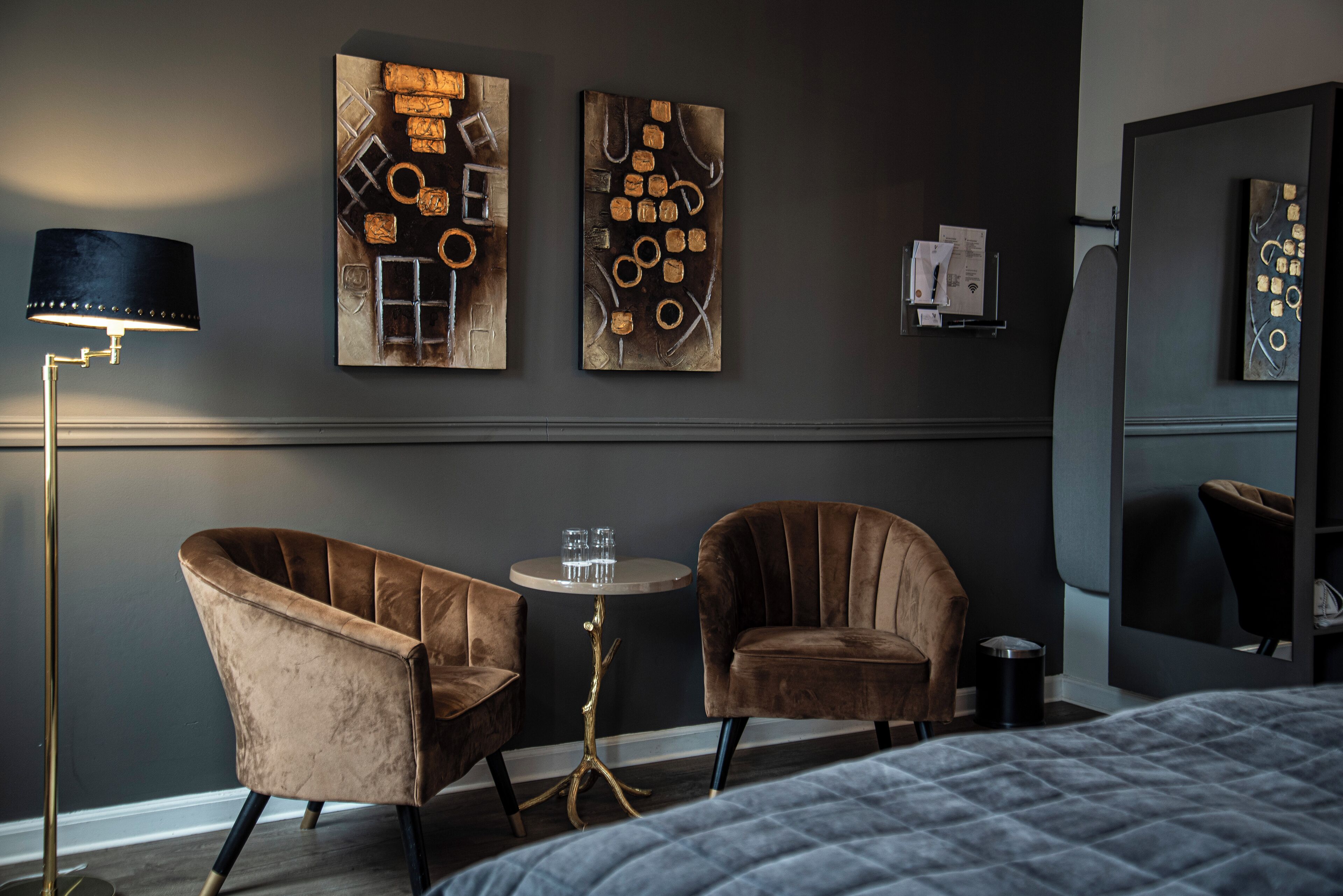 A modern hotel room with two velvet armchairs, a small round table with glasses, a floor lamp, and abstract artwork on the walls.