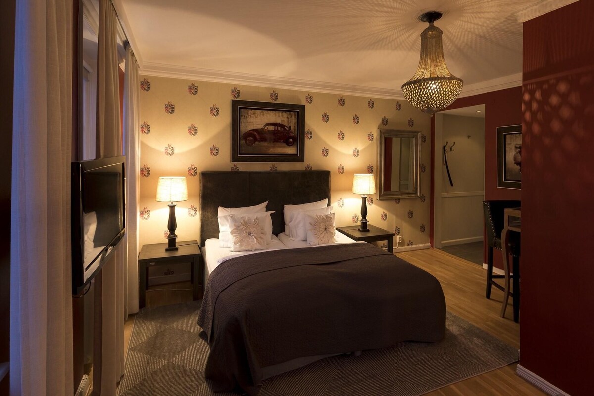 Habitación de hotel con cama, dos lámparas de noche, televisor, un adornado panel de pared y un candelabro.