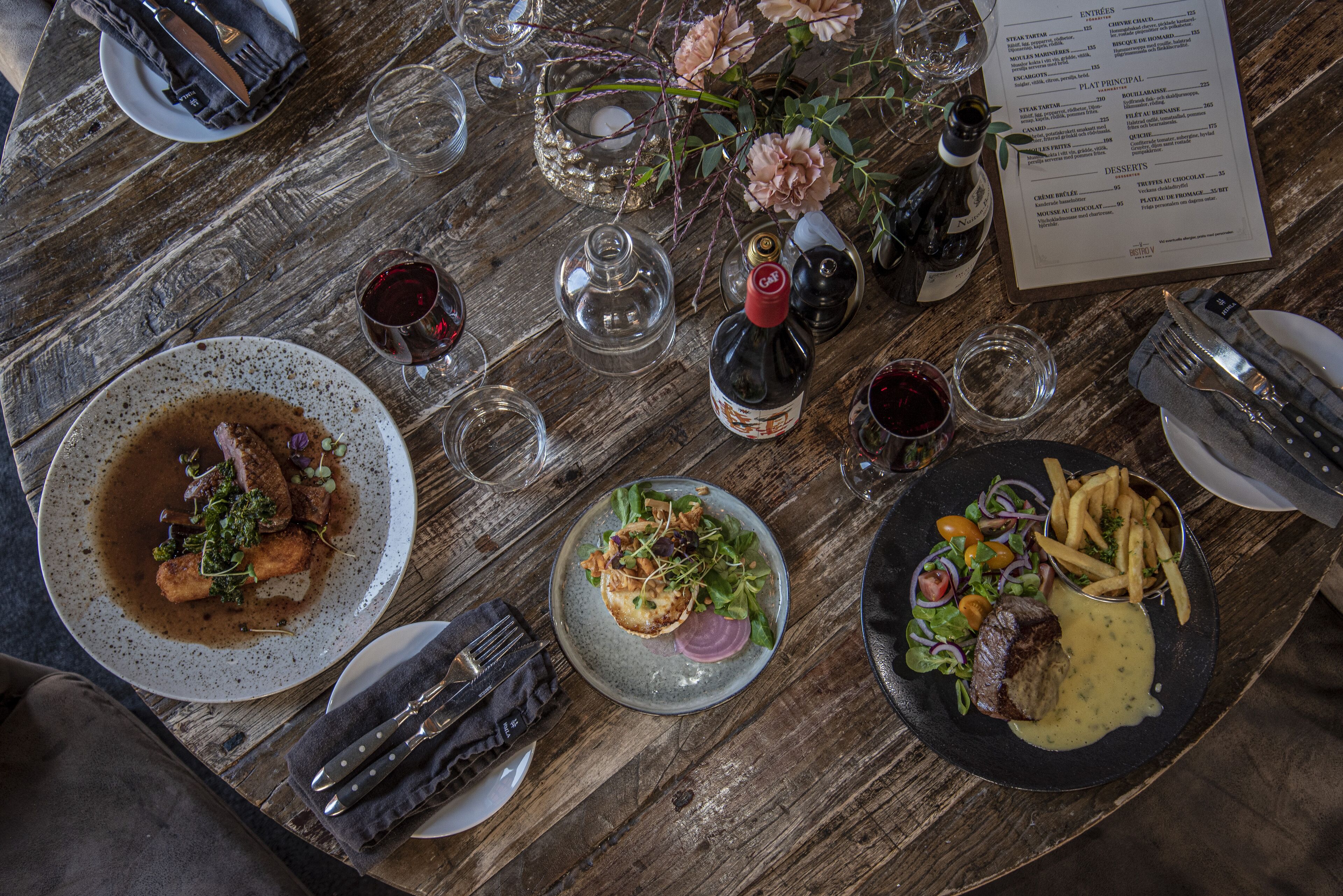 A dining table set with plates of food, wine glasses, a menu, and cutlery.