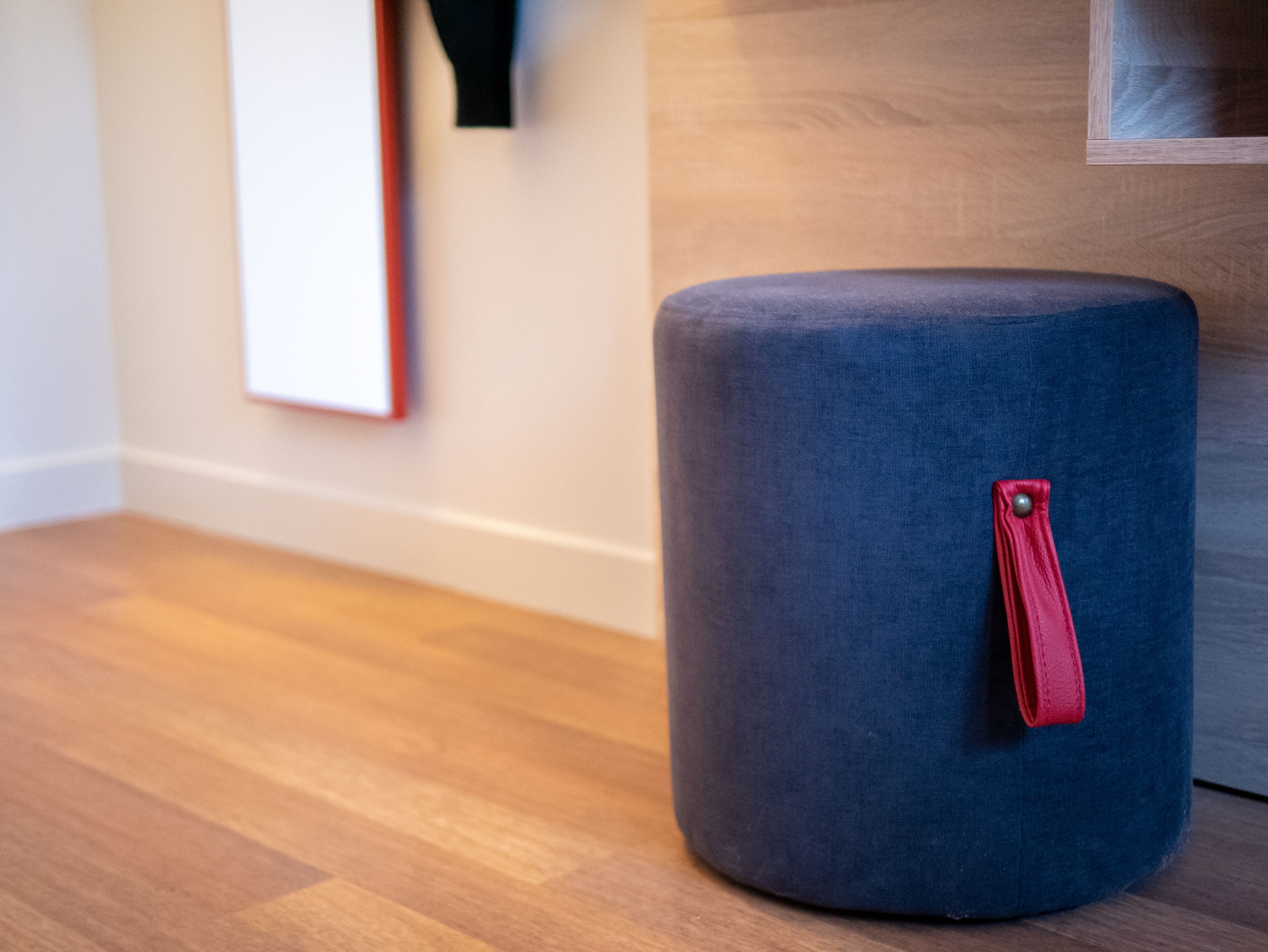 Un otoman redondo, azul oscuro, con una correa roja y un botón plateado, sobre un piso de madera junto a una pared con un cuadro enmarcado.