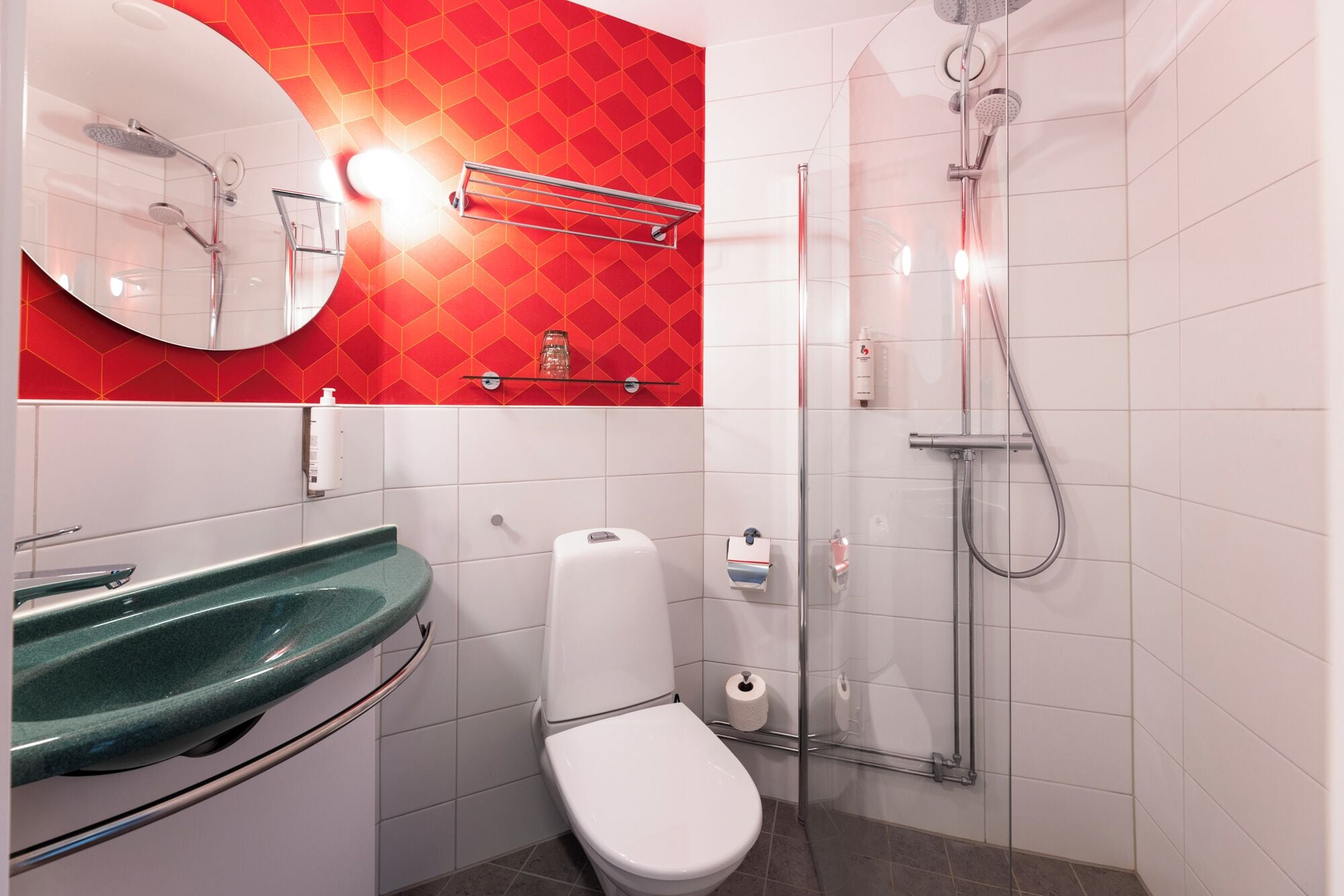 A modern bathroom with a white toilet, a green sink, a glass shower enclosure, and a red patterned wall.