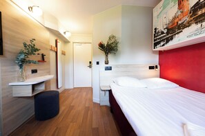 Une chambre d’hôtel moderne avec un plancher en bois, un lit avec une literie blanche, une petite table de chevet, un miroir et un luminaire fixé au mur.