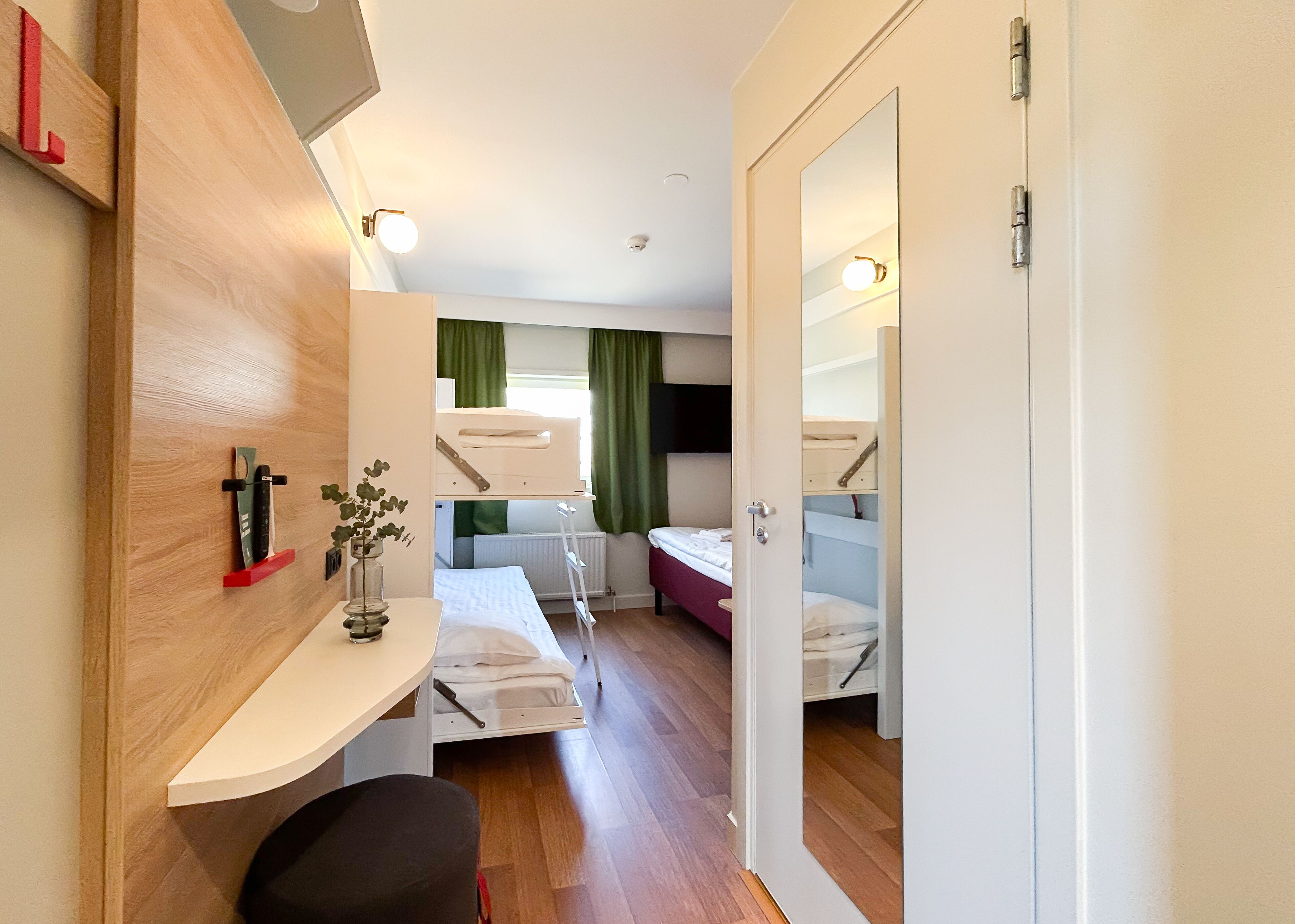 A compact hotel room with a bunk bed, a desk with a chair, a small table, and a mirror.