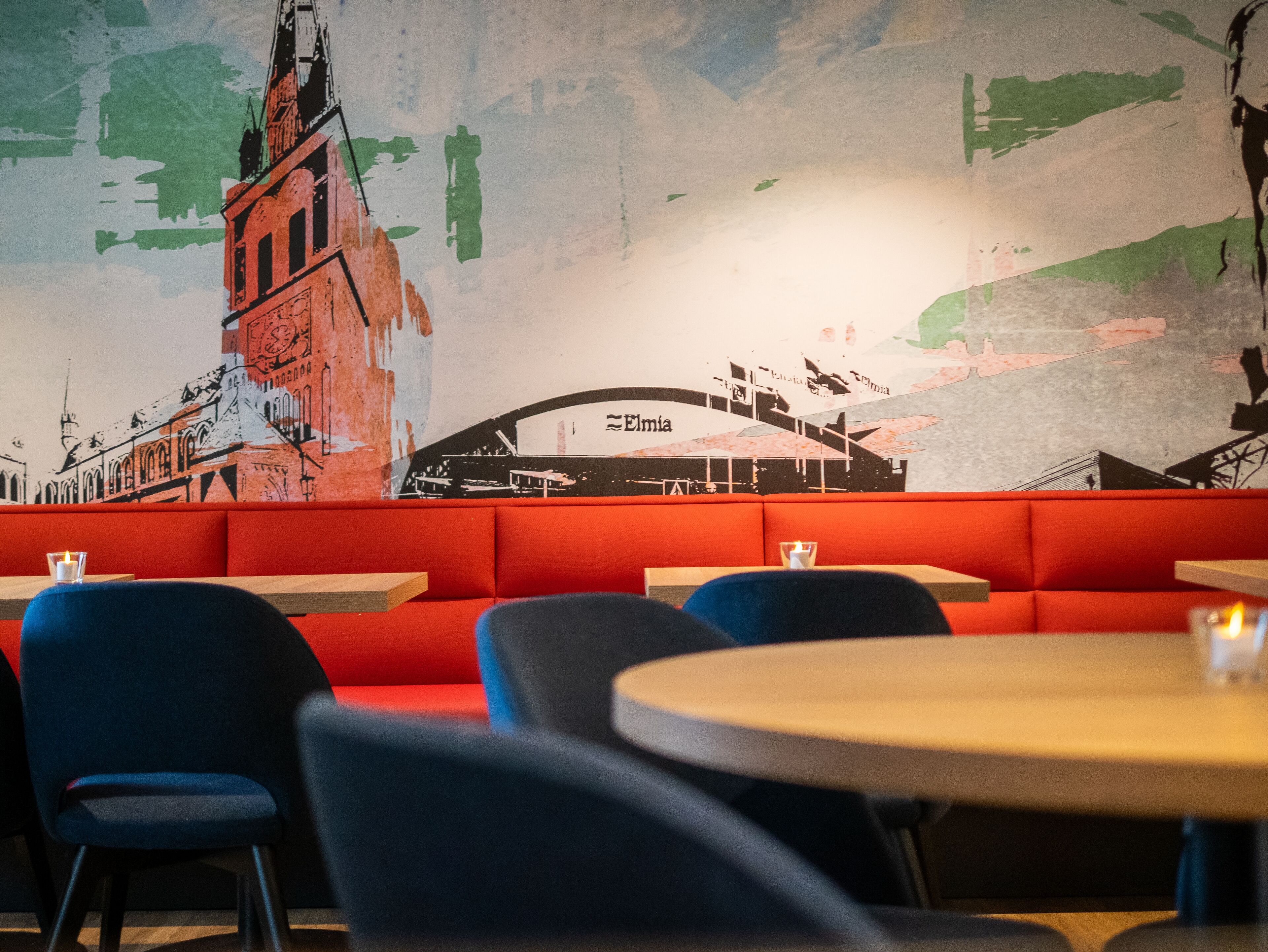 En restaurang med en inredning som kännetecknas av röda bås, runda bord och en väggmålning som föreställer en stadsbild.