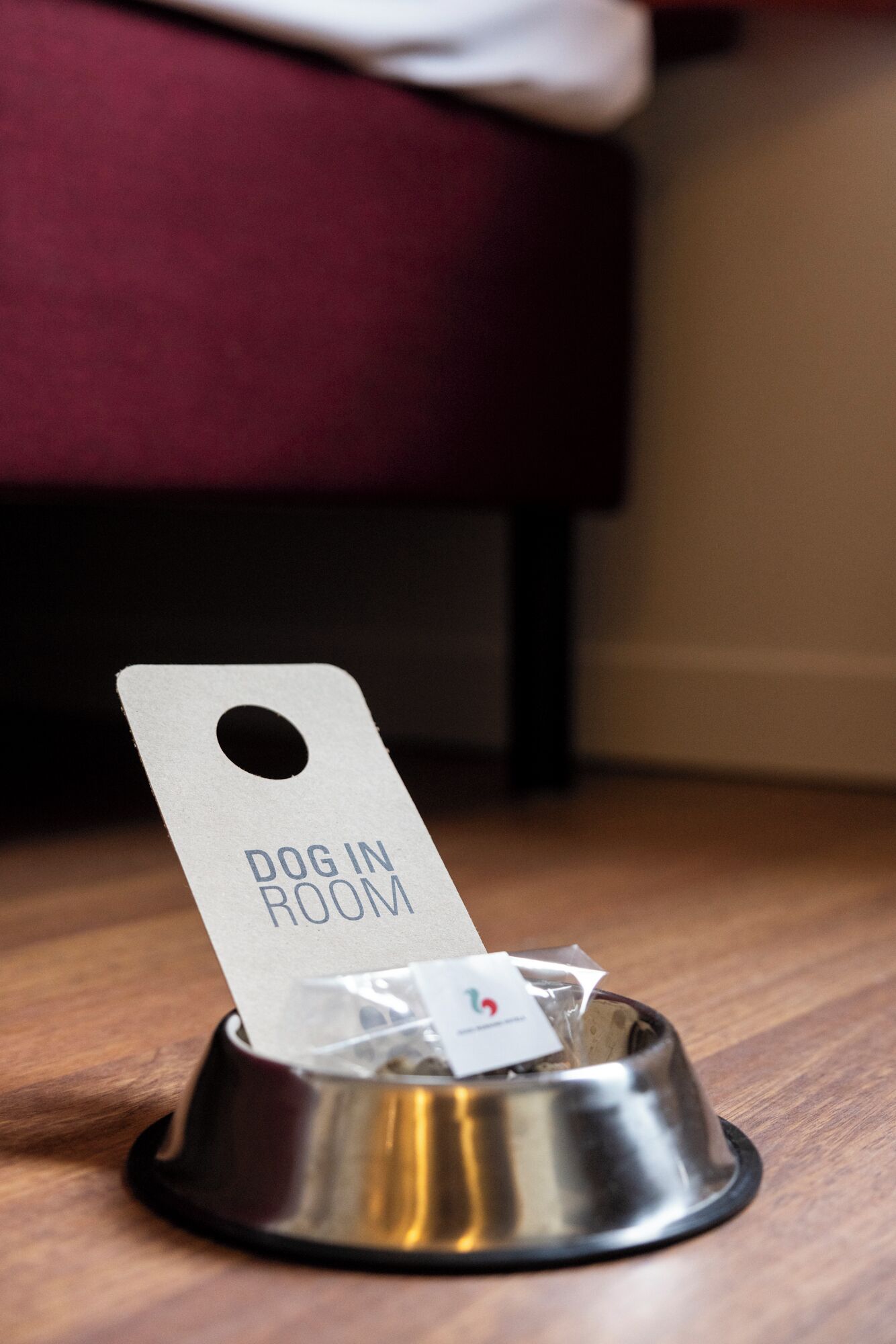 A metal dog bowl with a tag that reads "DOG IN ROOM" on a wooden floor.