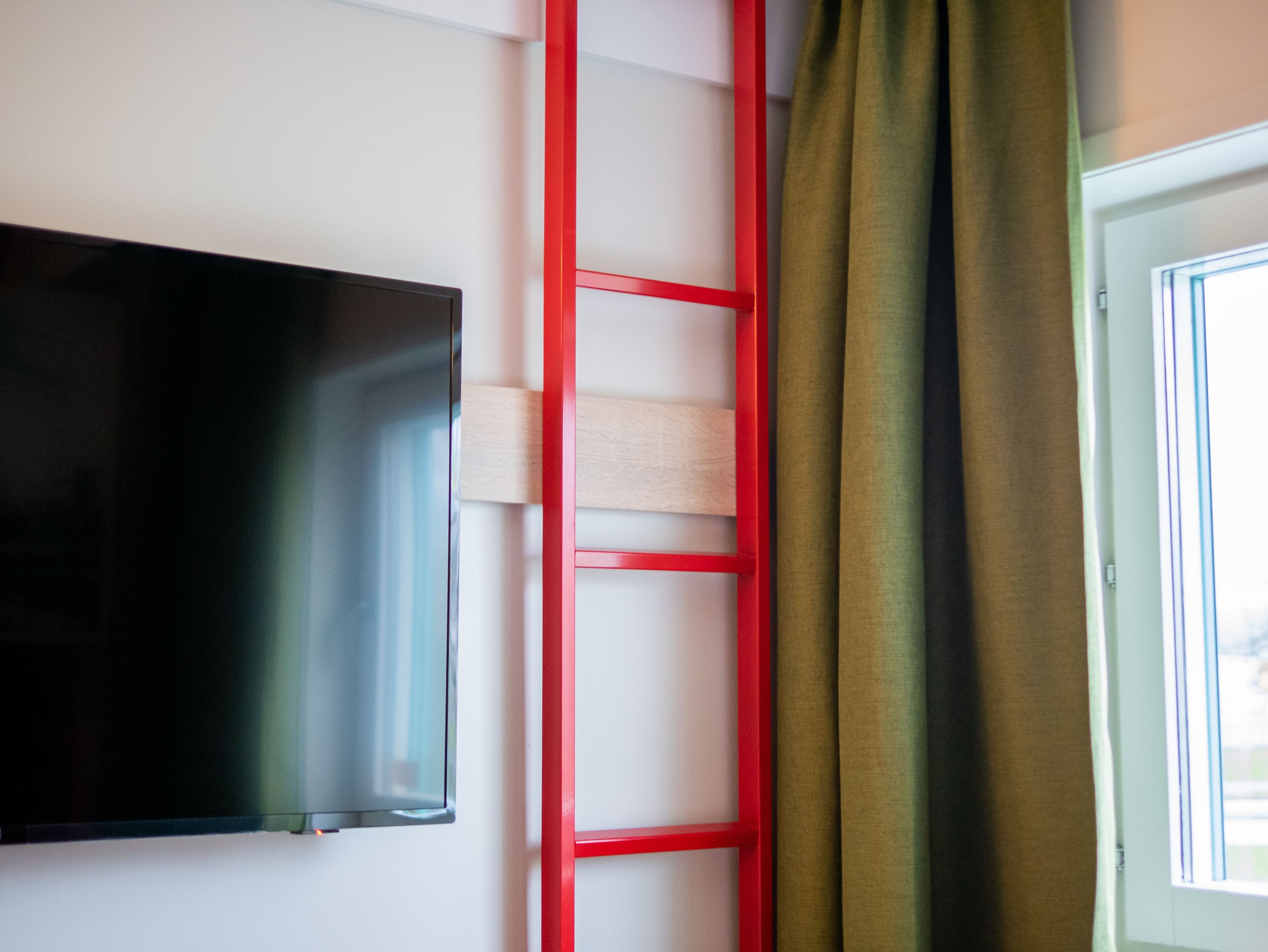 Una habitación con una televisión de pantalla plana en la pared, una escalera roja apoyada en la pared y una ventana con cortinas.