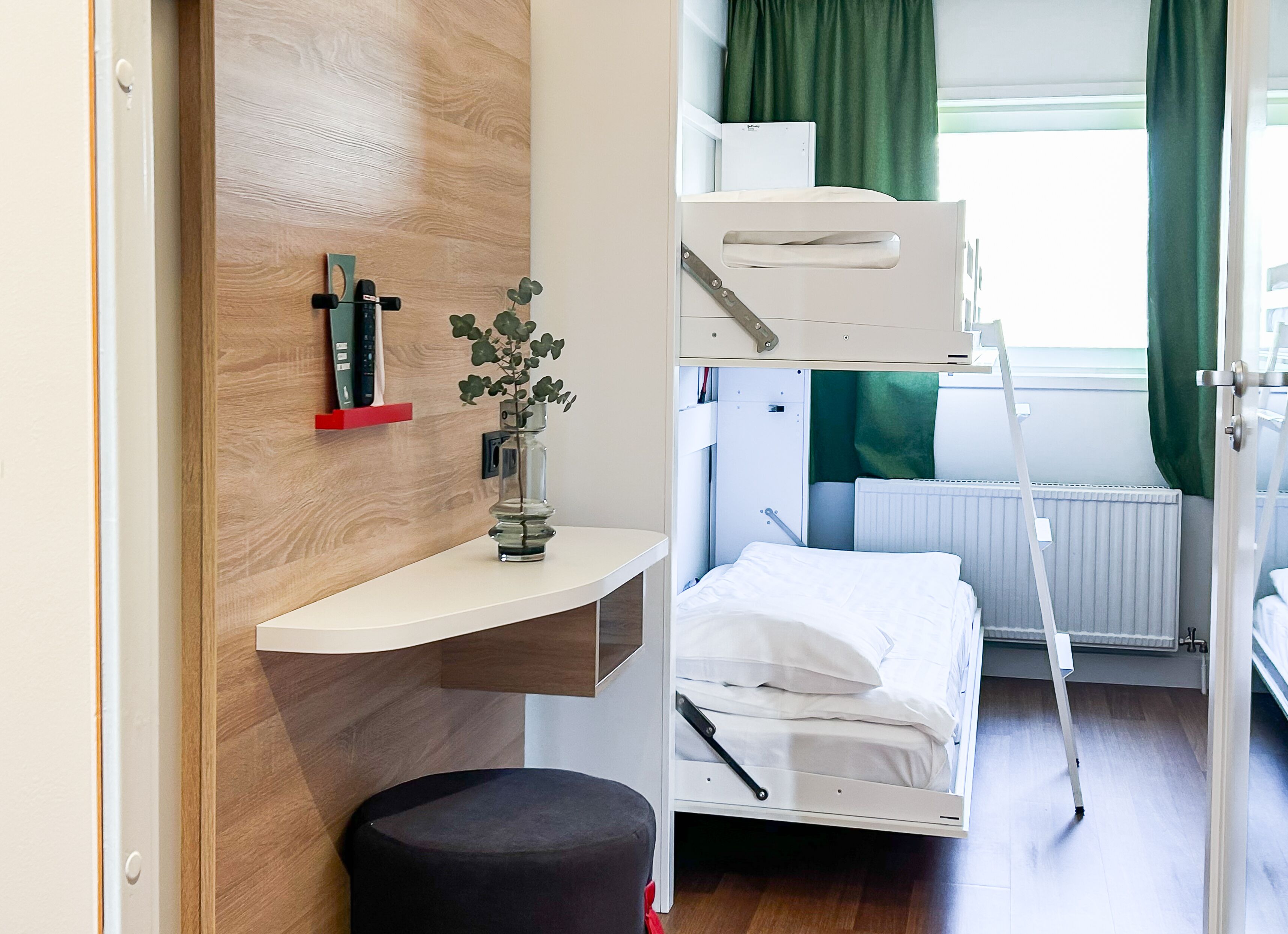 A bunk bed with a desk and a small stool, a radiator, and a window with curtains.