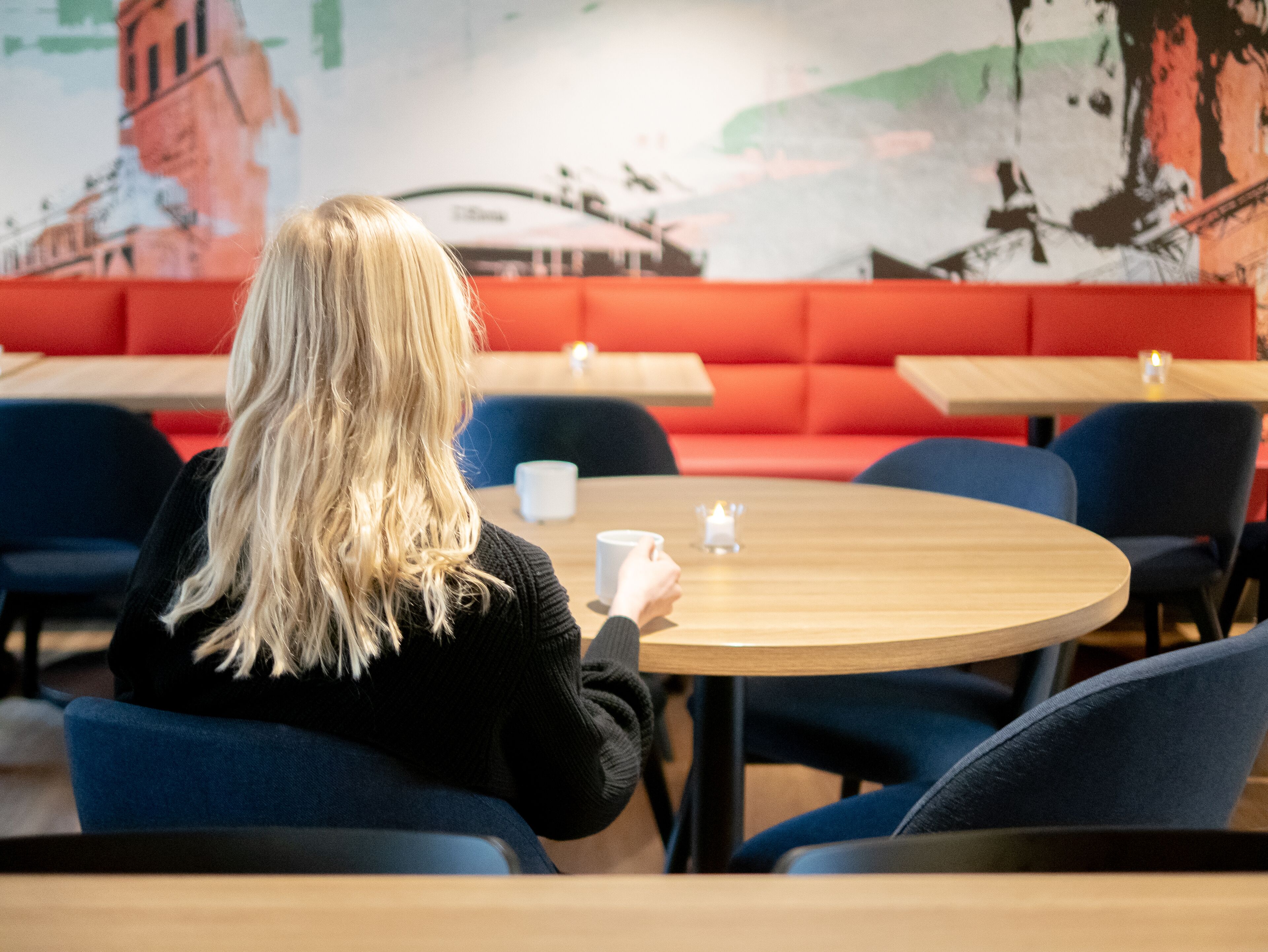 Een persoon zit aan een ronde tafel in een café met rode stoelen en een muurschildering op de achtergrond.