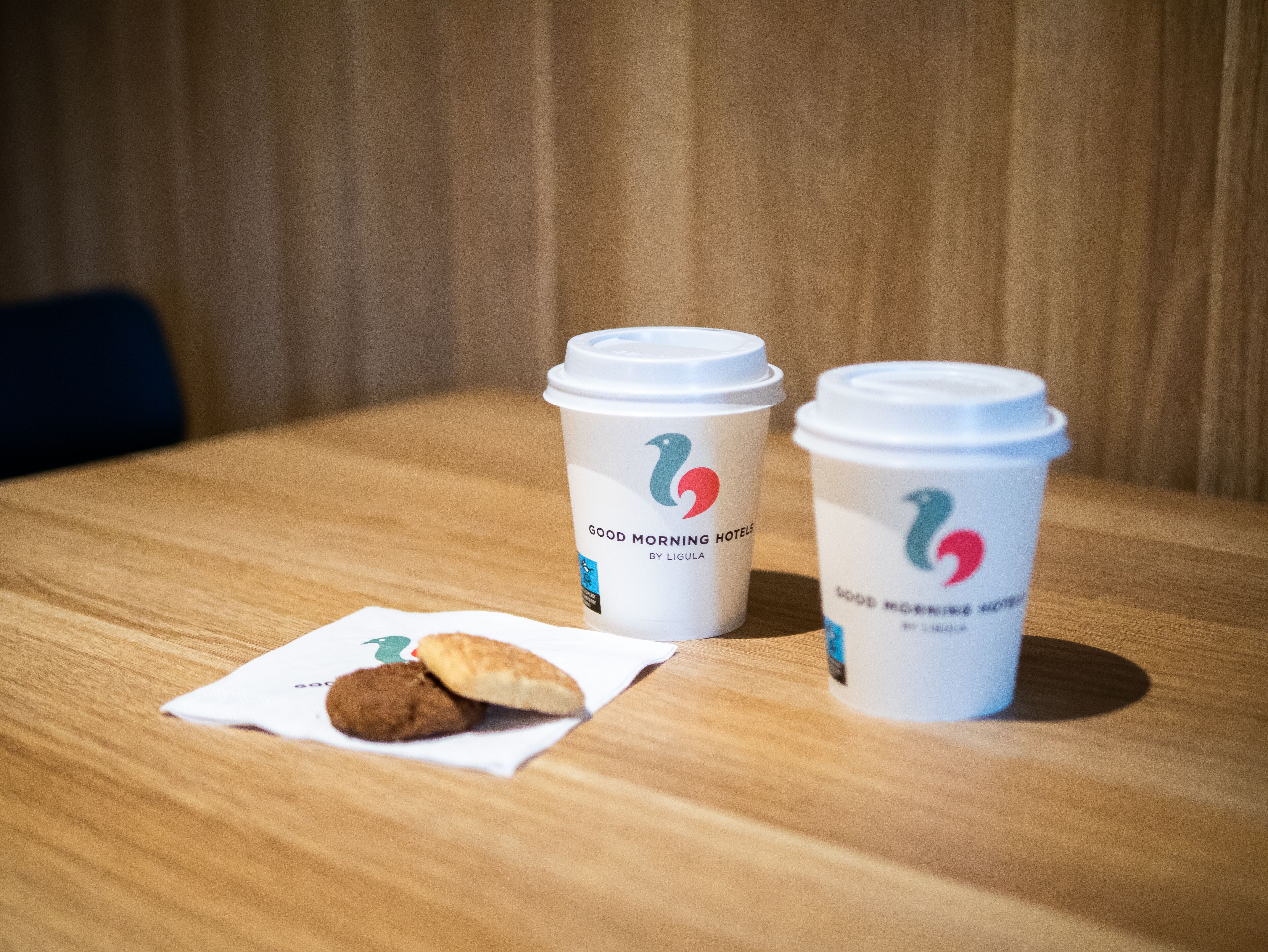 Two disposable coffee cups with lids on a wooden table, one with a cookie.